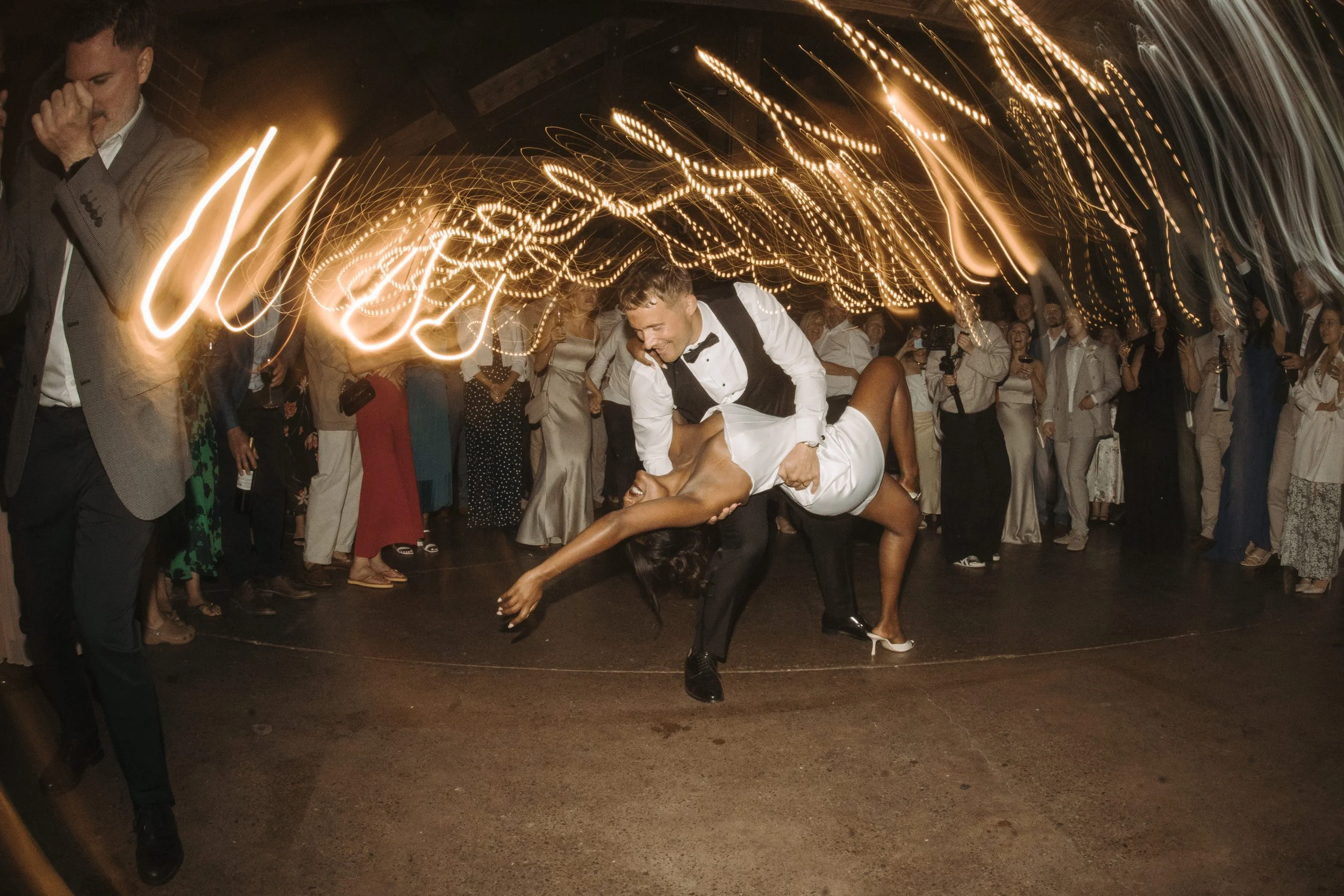 Couple dancing closely at night with guests watching, illuminated by string lights overhead.