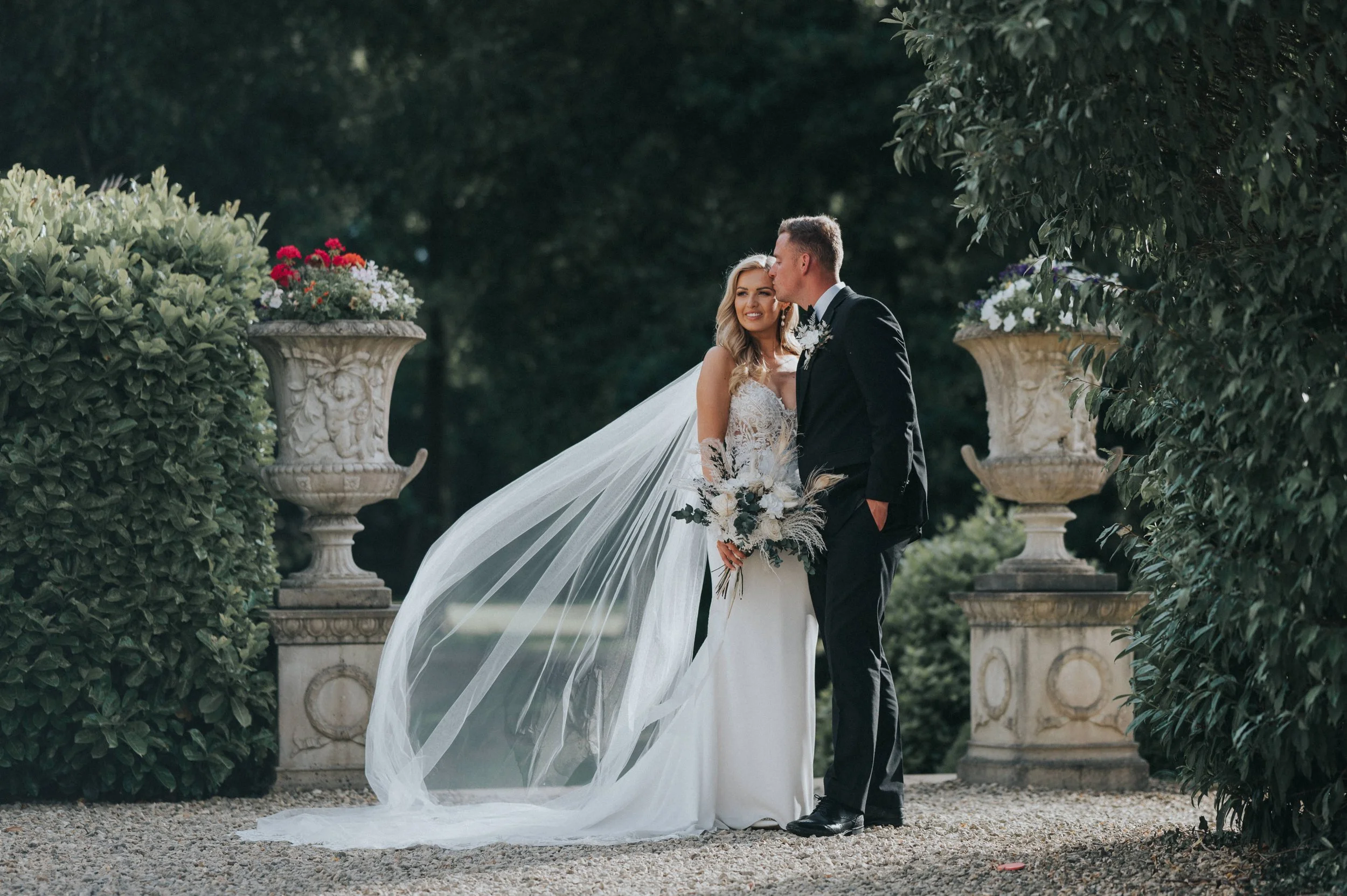 A bride and groom standing close together outdoors during a wedding, with the groom kissing the bride on her forehead. The bride is wearing a white wedding dress with lace and a long veil, holding a bouquet of flowers. They are surrounded by lush gre