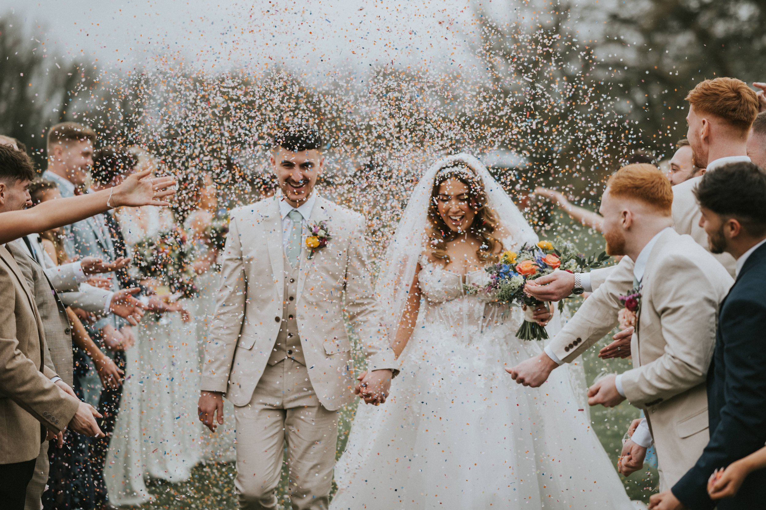 Bride and groom holding hands walking through a colorful confetti celebration, surrounded by friends and family at an outdoor wedding.