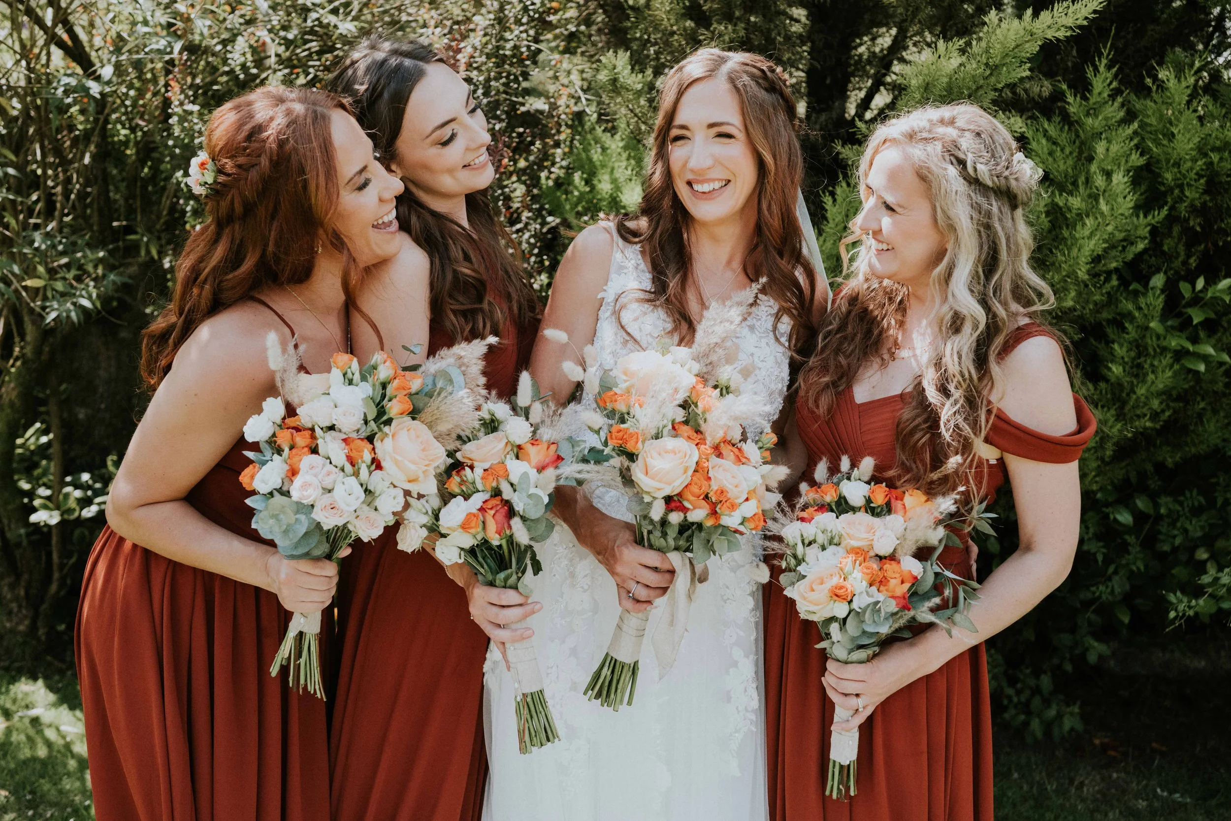 A group of women at a wedding, including the bride in a white dress and bridesmaids in matching burgundy dresses, holding bouquets of flowers outdoors.