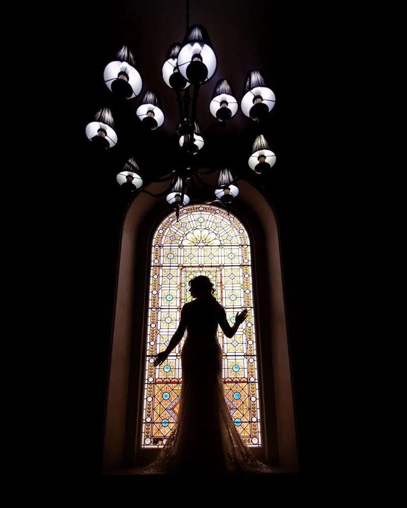 Silhouette of a woman in a long dress standing in front of a stained glass window, with a chandelier hanging above.