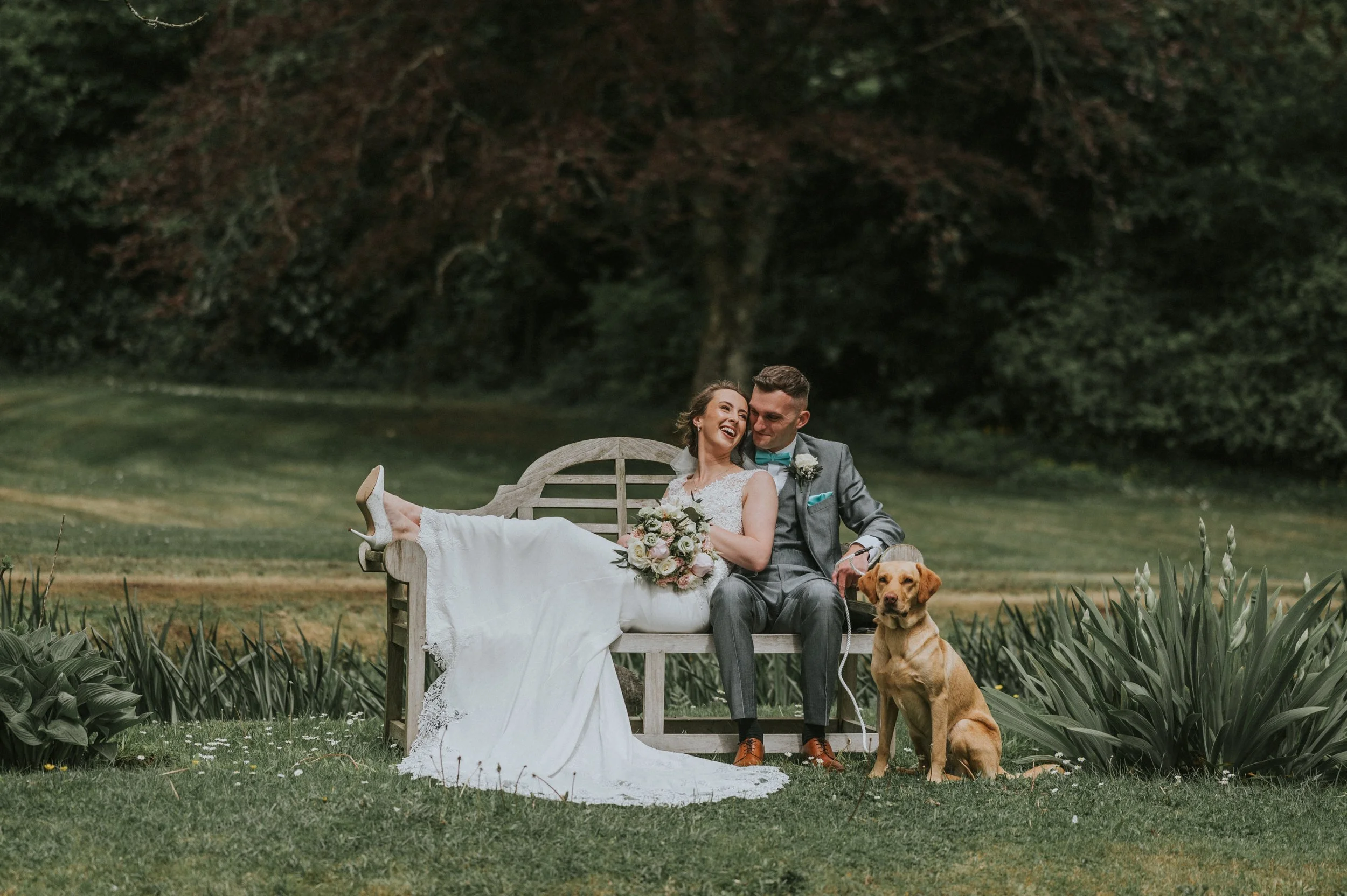 A bride and groom sitting on a wooden bench outdoors, with the bride in a white wedding dress and holding a bouquet, and the groom in a gray suit with a teal bow tie, both smiling and looking at each other. A golden retriever dog is sitting in front 