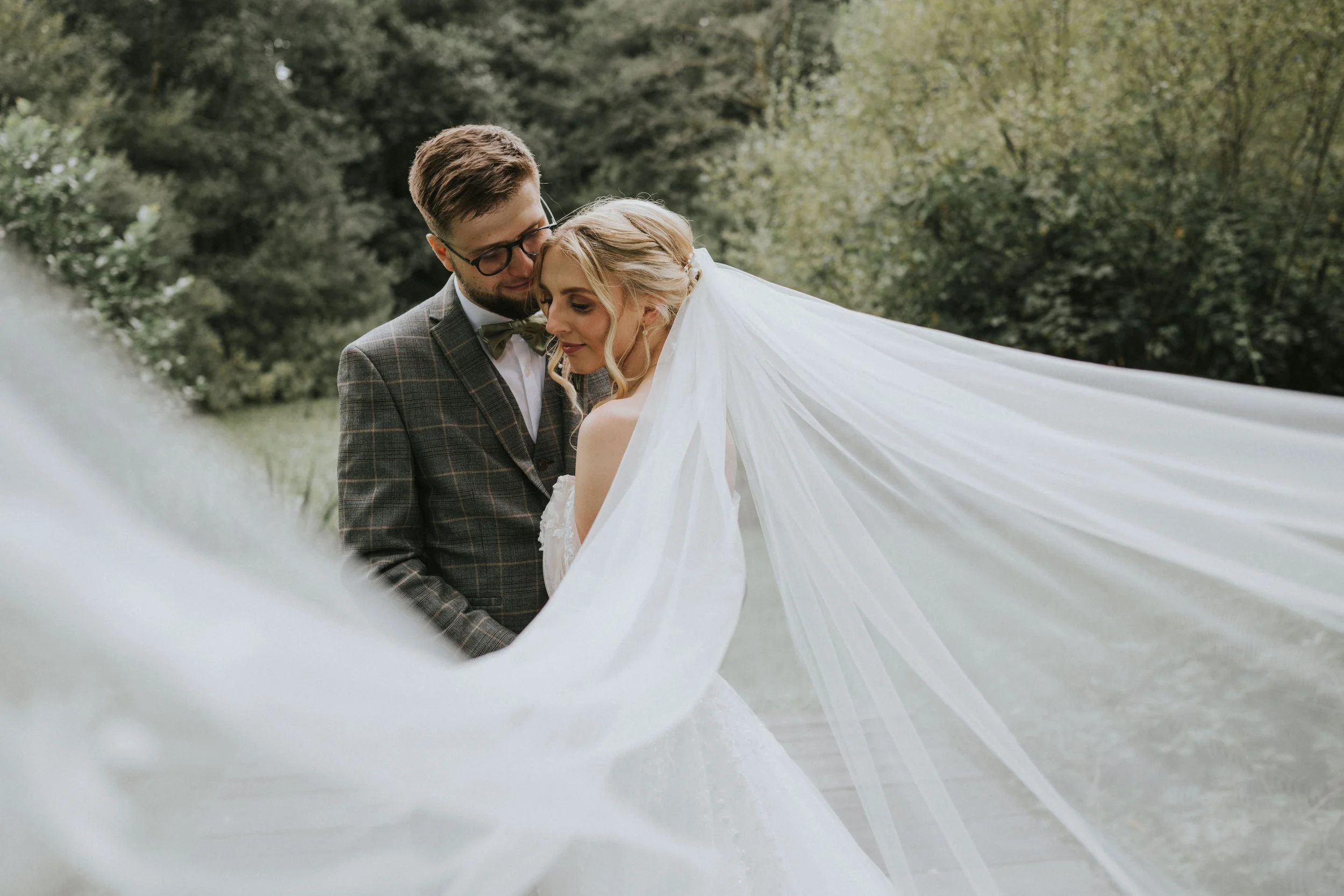 A bride and groom embracing outdoors, with the bride wearing a wedding dress and a long veil, and the groom in a suit, surrounded by greenery.