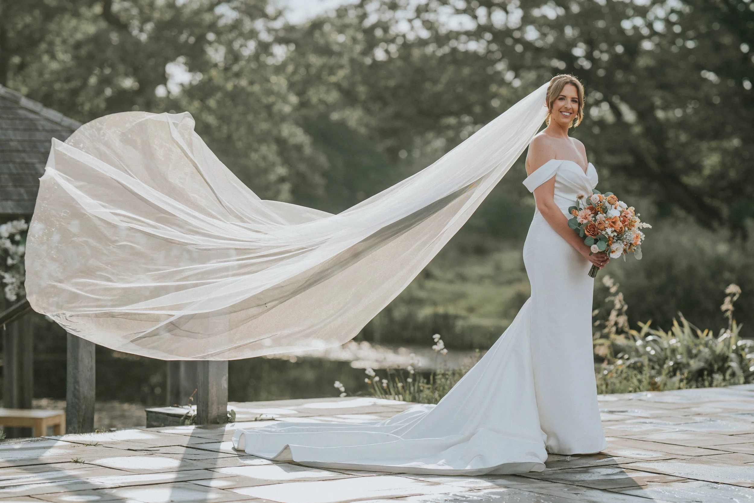 A bride in a white off-shoulder wedding gown holding a bouquet of flowers, standing outdoors with a long veil flowing behind her, smiling.