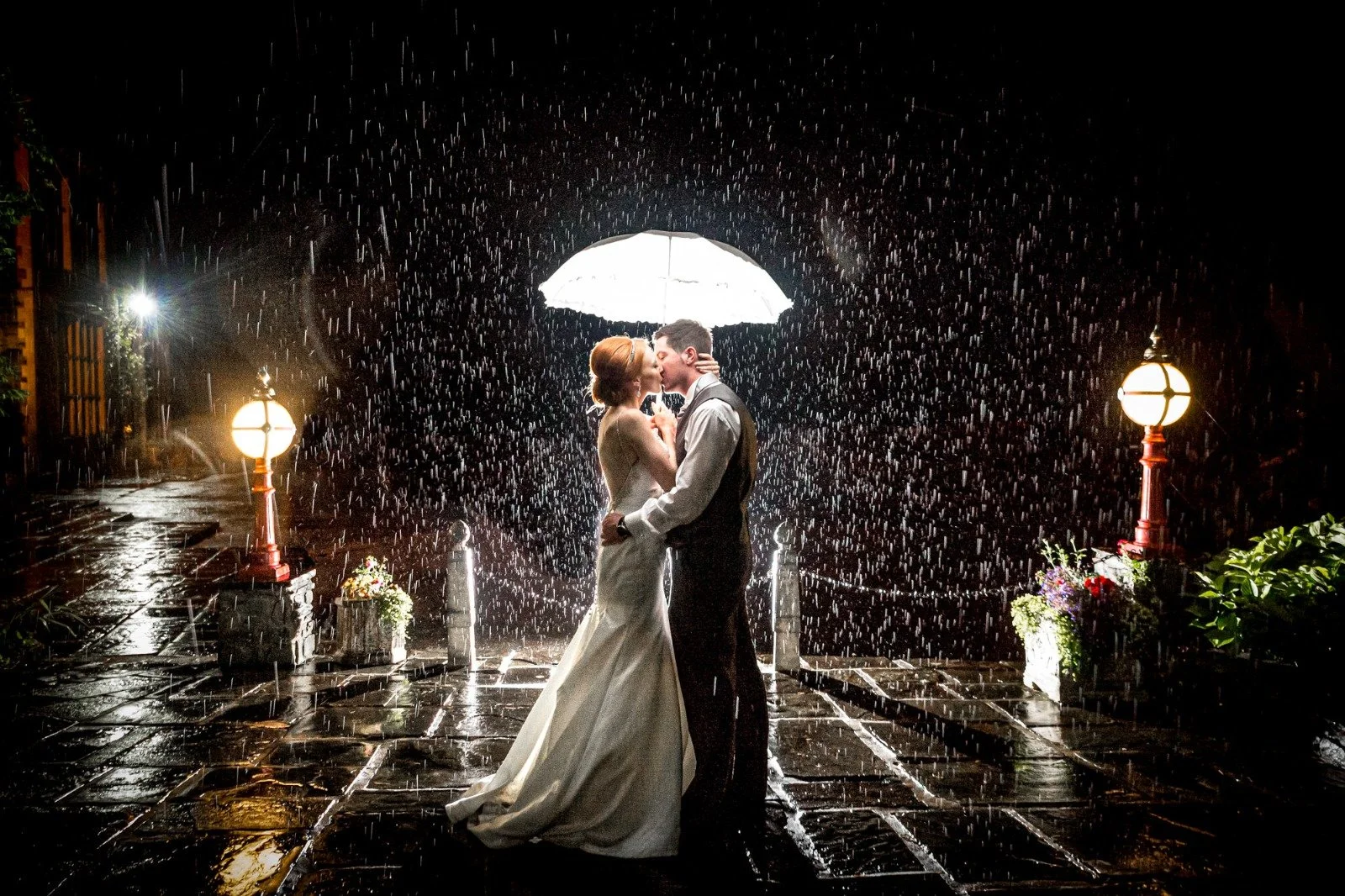 A bride and groom kiss under an umbrella in the rain at night, illuminated by two street lamps on either side, with flowers nearby.
