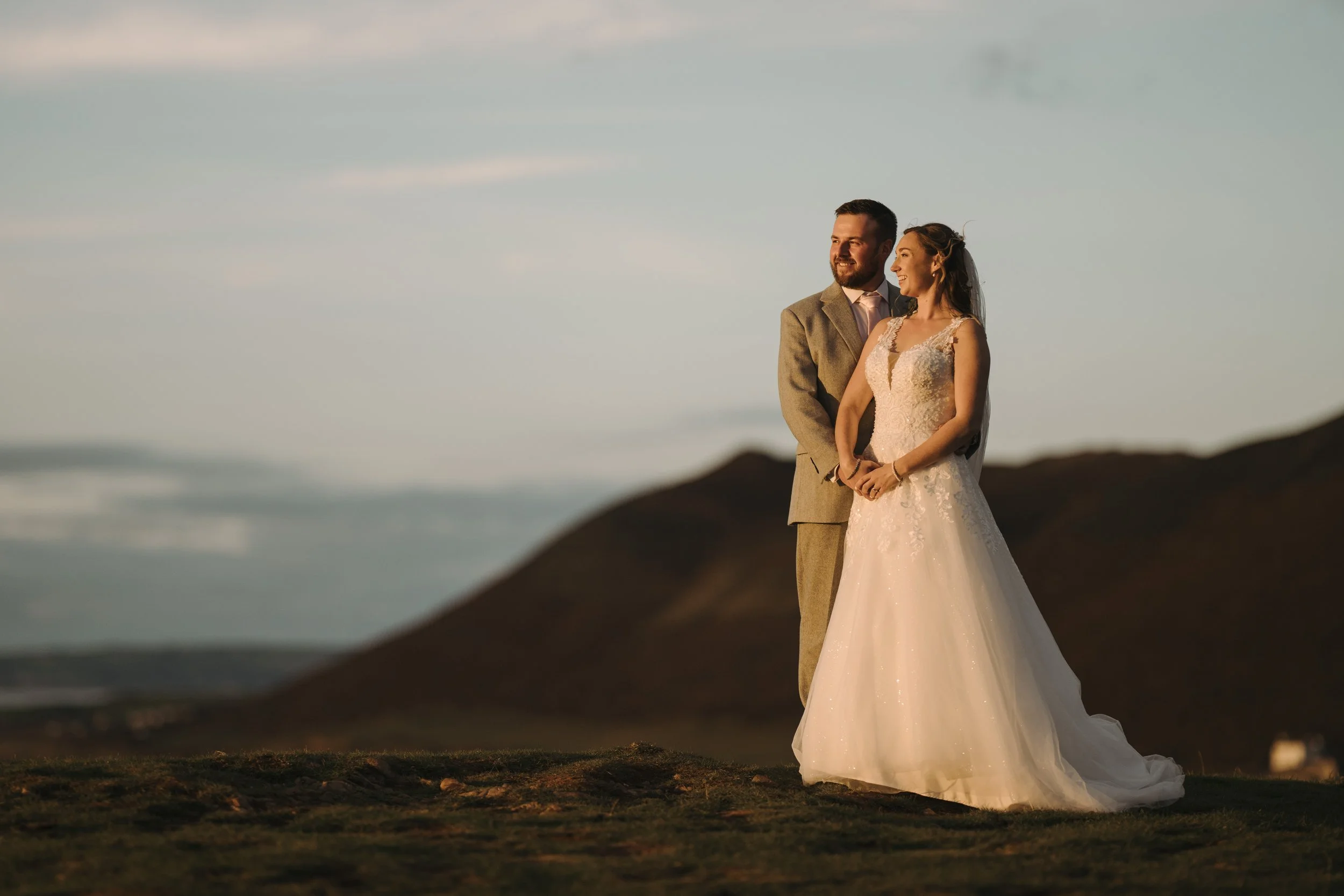 A couple in wedding attire standing outdoors at sunset. The bride is wearing a white wedding dress with lace details, and the groom is wearing a grey suit with a white shirt and tie. They are smiling and holding hands, with a blurred natural landscap