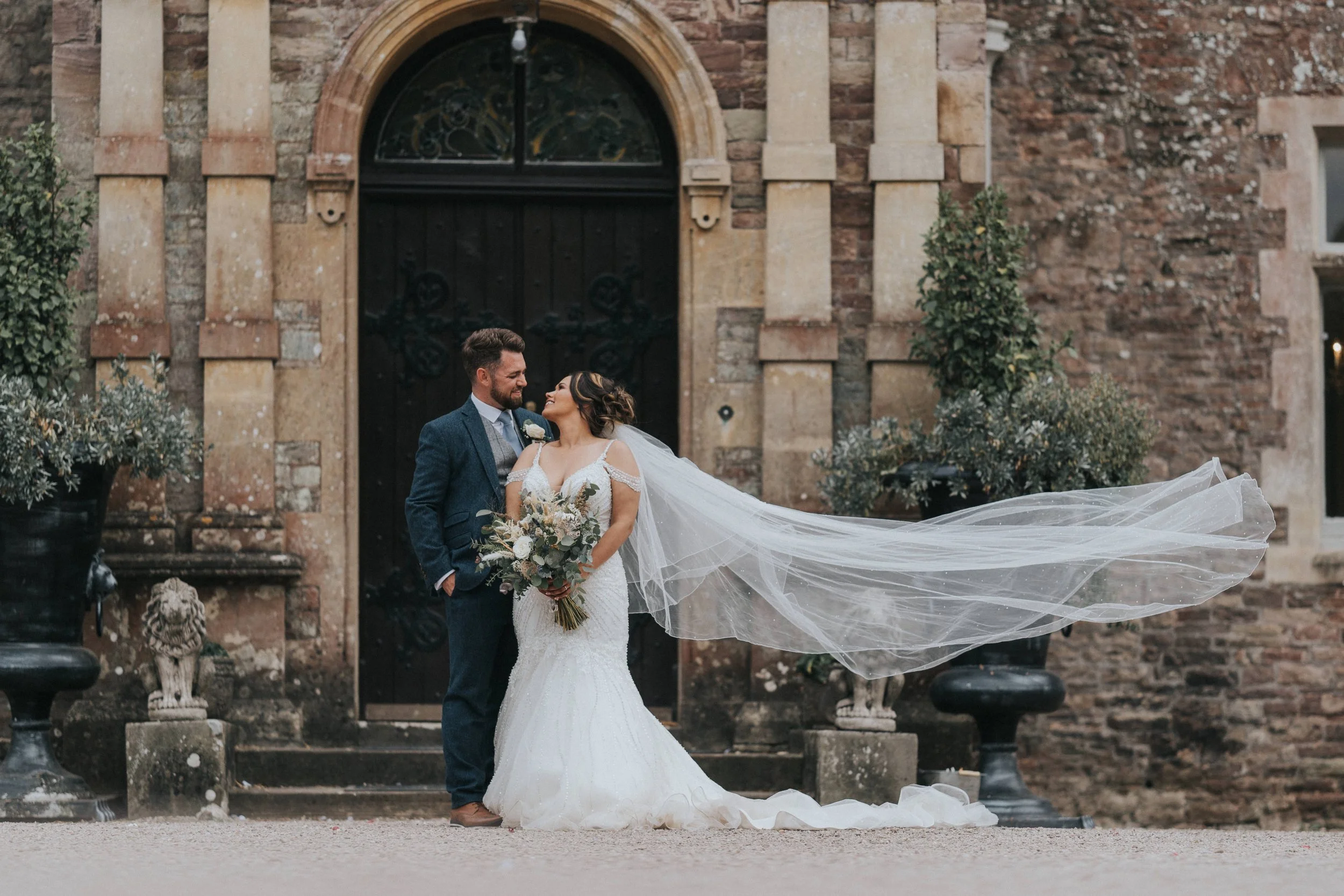A bride and groom stand close together outside in front of a brick building, smiling at each other. The bride wears a white wedding gown with a long veil flowing in the breeze, and holds a bouquet of flowers. The groom is dressed in a blue suit. Ther