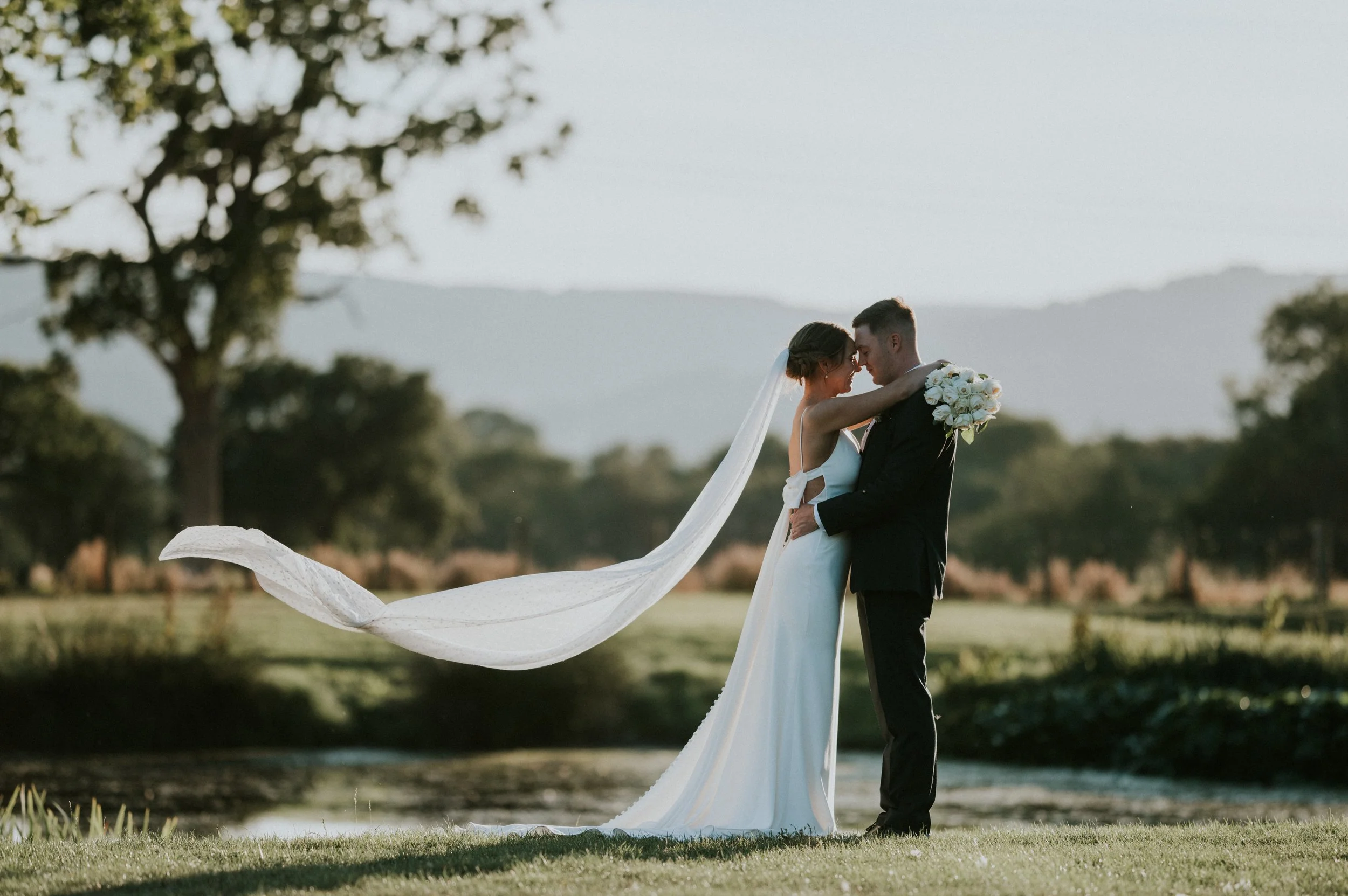A bride and groom embracing outdoors near trees and mountains, with the bride holding a bouquet of white roses and wearing a long veil.