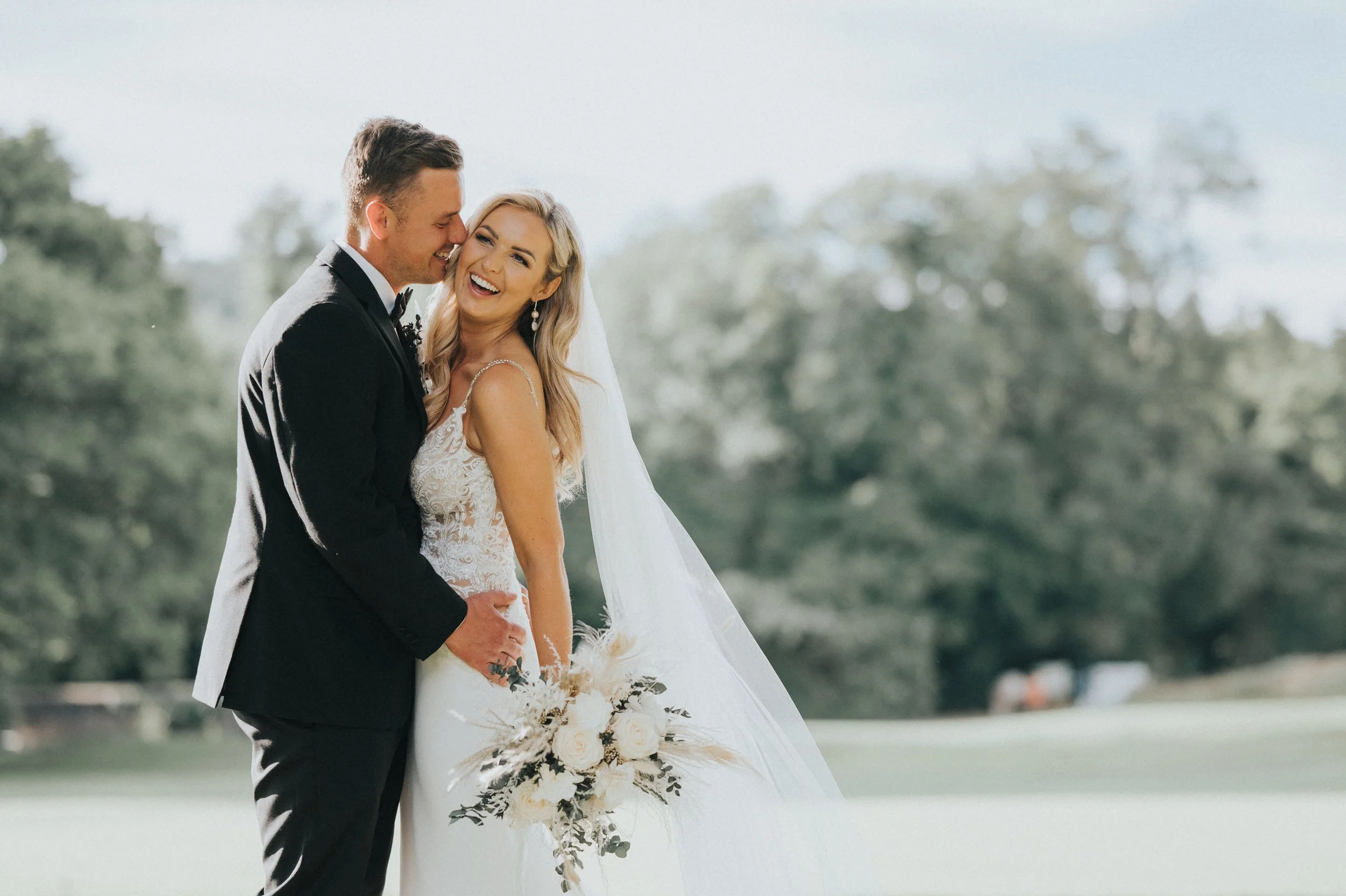 A happy bride and groom on their wedding day, standing outdoors with trees in the background. The bride is smiling and holding a bouquet of white flowers, while the groom is leaning in close, smiling.