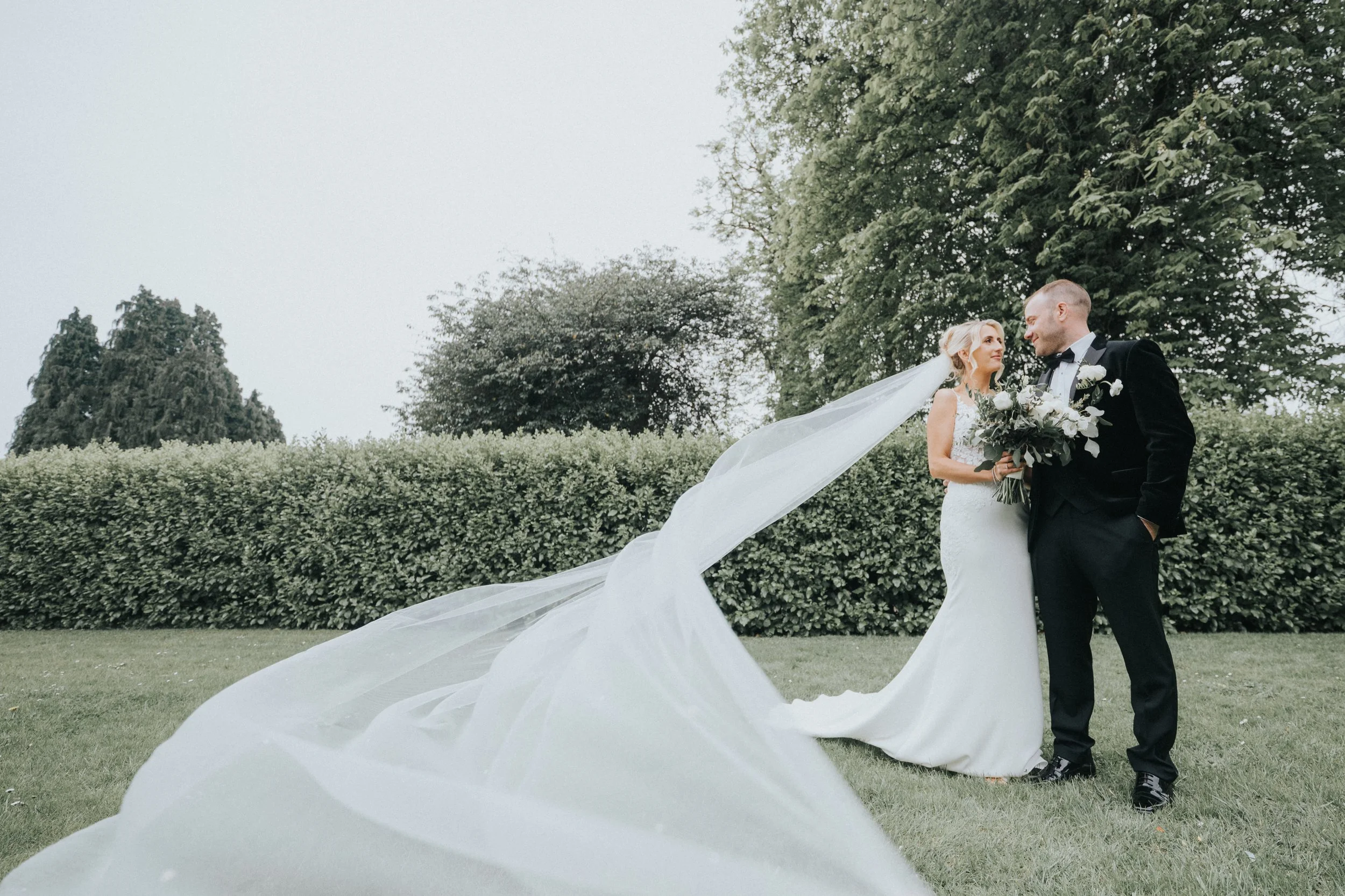 Bride and groom standing outdoors with greenery, the bride holding a bouquet, and the bride's veil flowing in the wind.