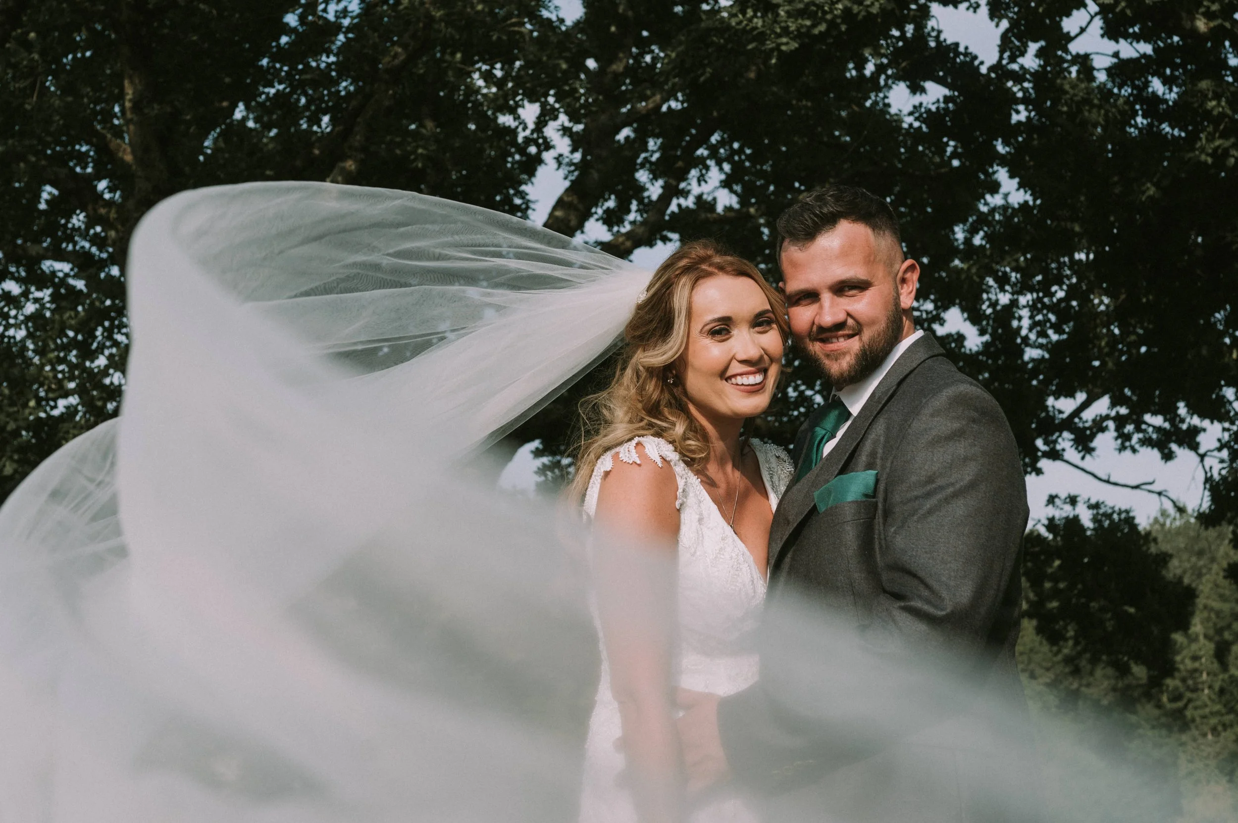A happy bride and groom smiling outdoors, with the bride wearing a white dress and veil, and the groom in a gray suit with a green tie, standing under a large tree.