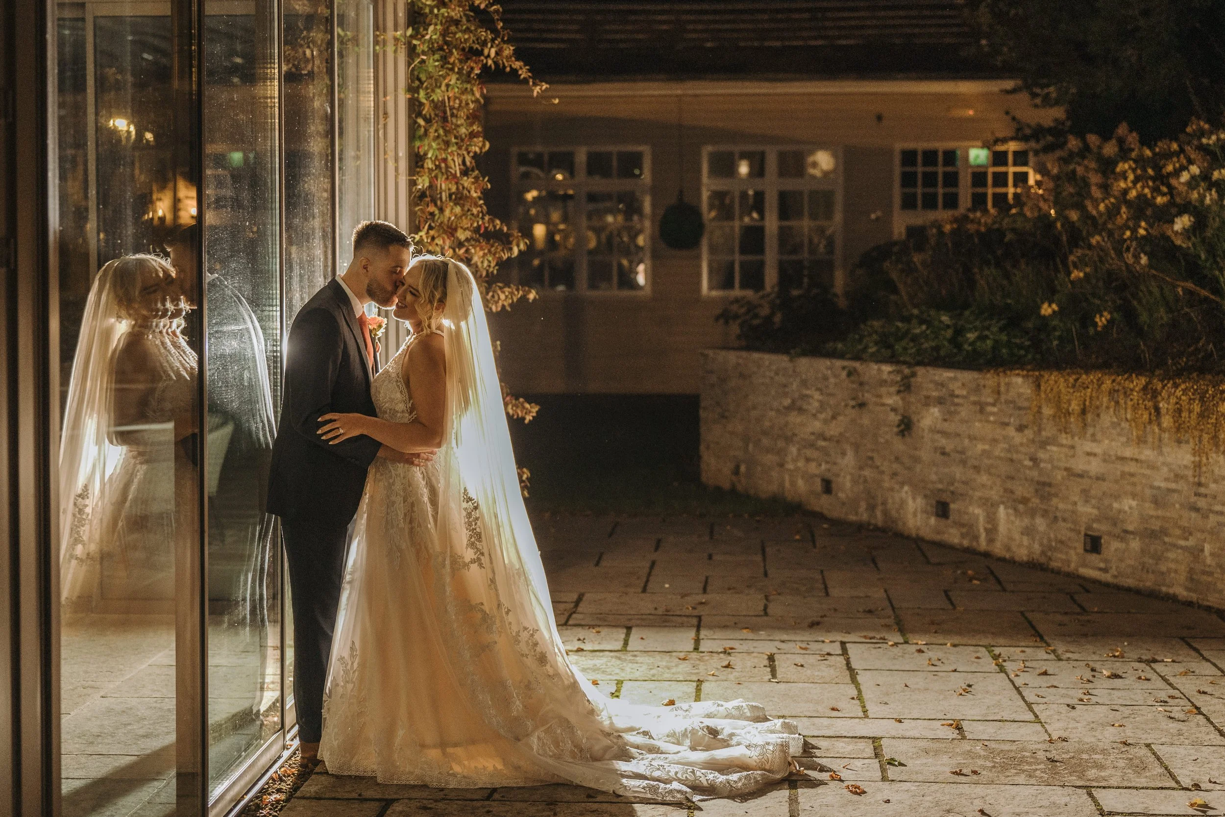 A bride and groom share a kiss outside a building illuminated by warm light at night, with the bride in a wedding gown and veil and the groom in a suit, reflected in glass windows.