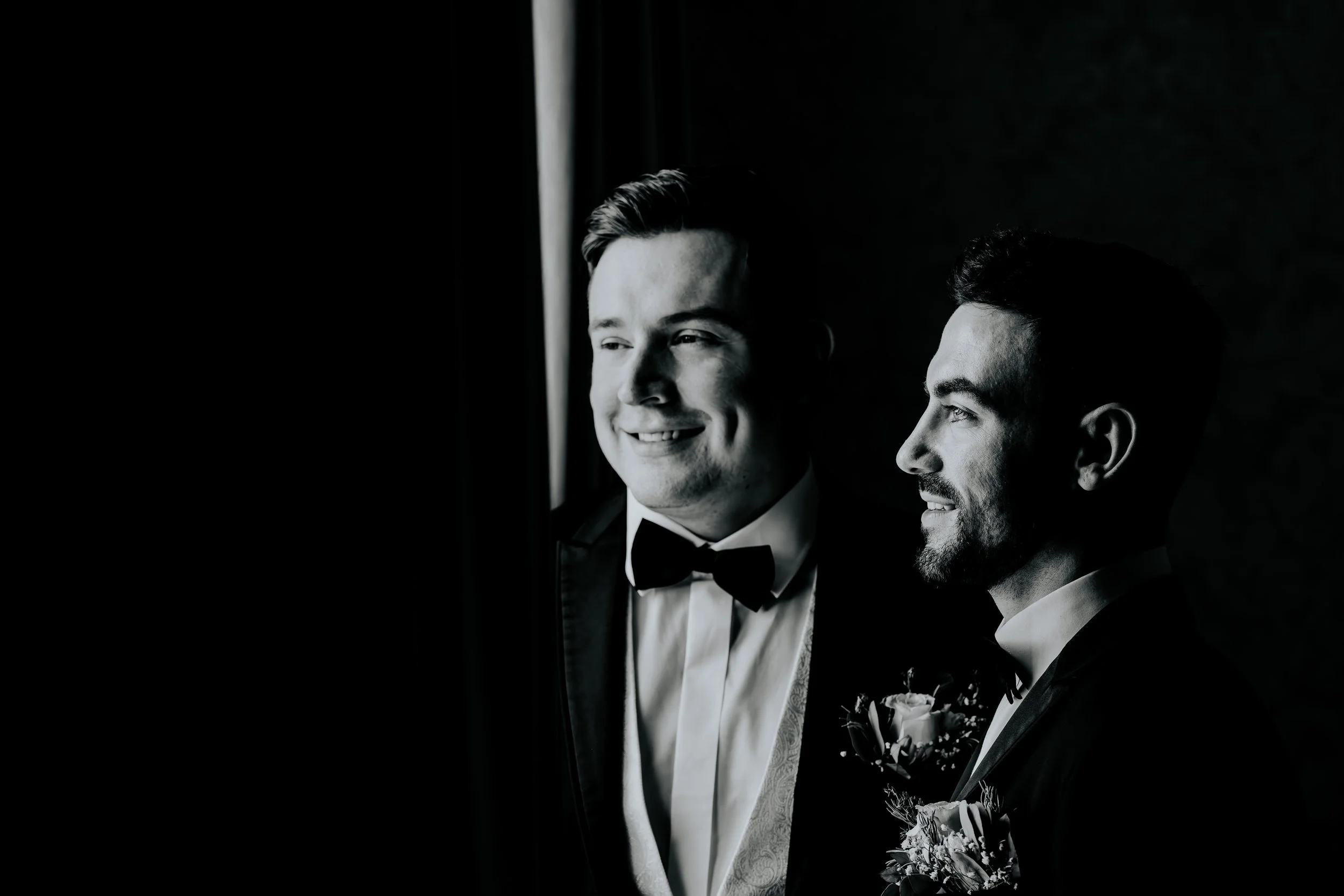 Two men dressed in tuxedos, standing near a window with dark curtains, smiling and looking out.