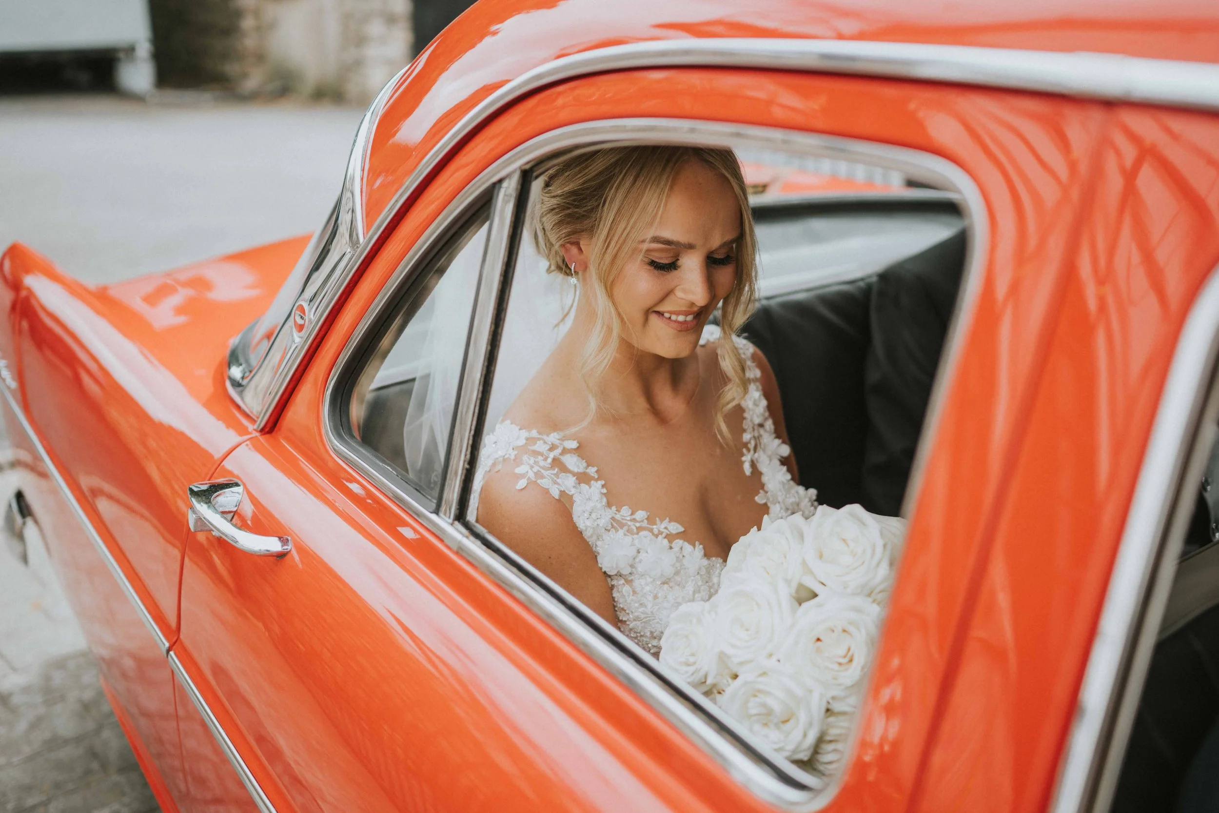 A bride sitting inside a vintage orange car, holding a bouquet of white roses, with a big smile.