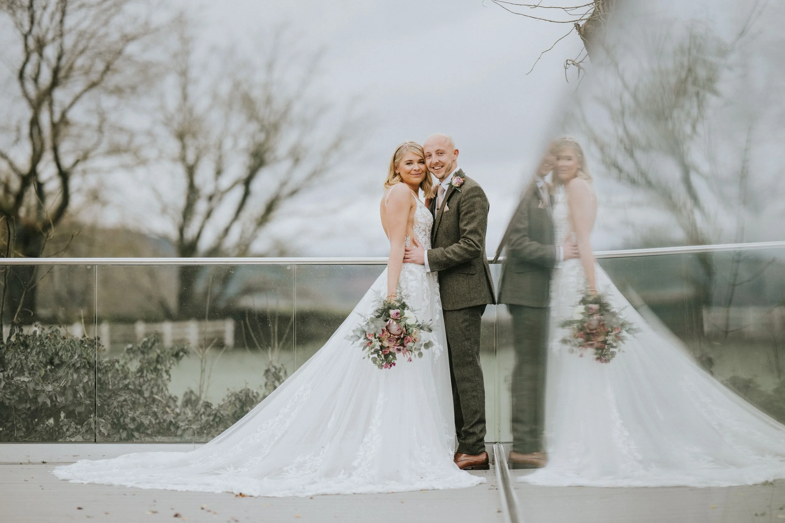 A bride and groom standing close together outdoors, with the bride holding a bouquet, reflected on a glass surface, in front of leafless trees and an overcast sky.
