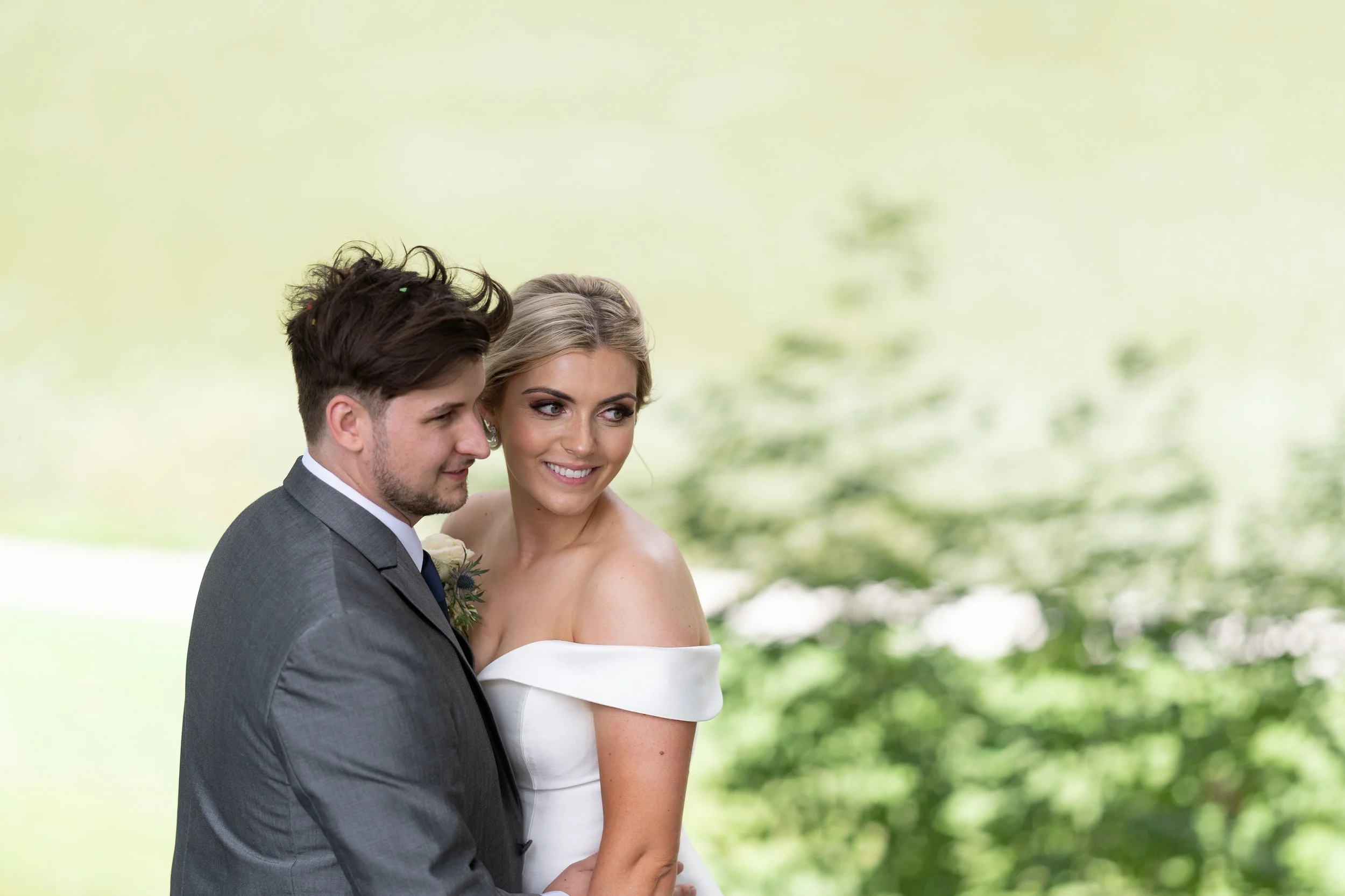 A bride and groom outdoors near a body of water, smiling with their heads close together. The groom wears a gray suit, and the bride wears an off-shoulder white wedding dress.