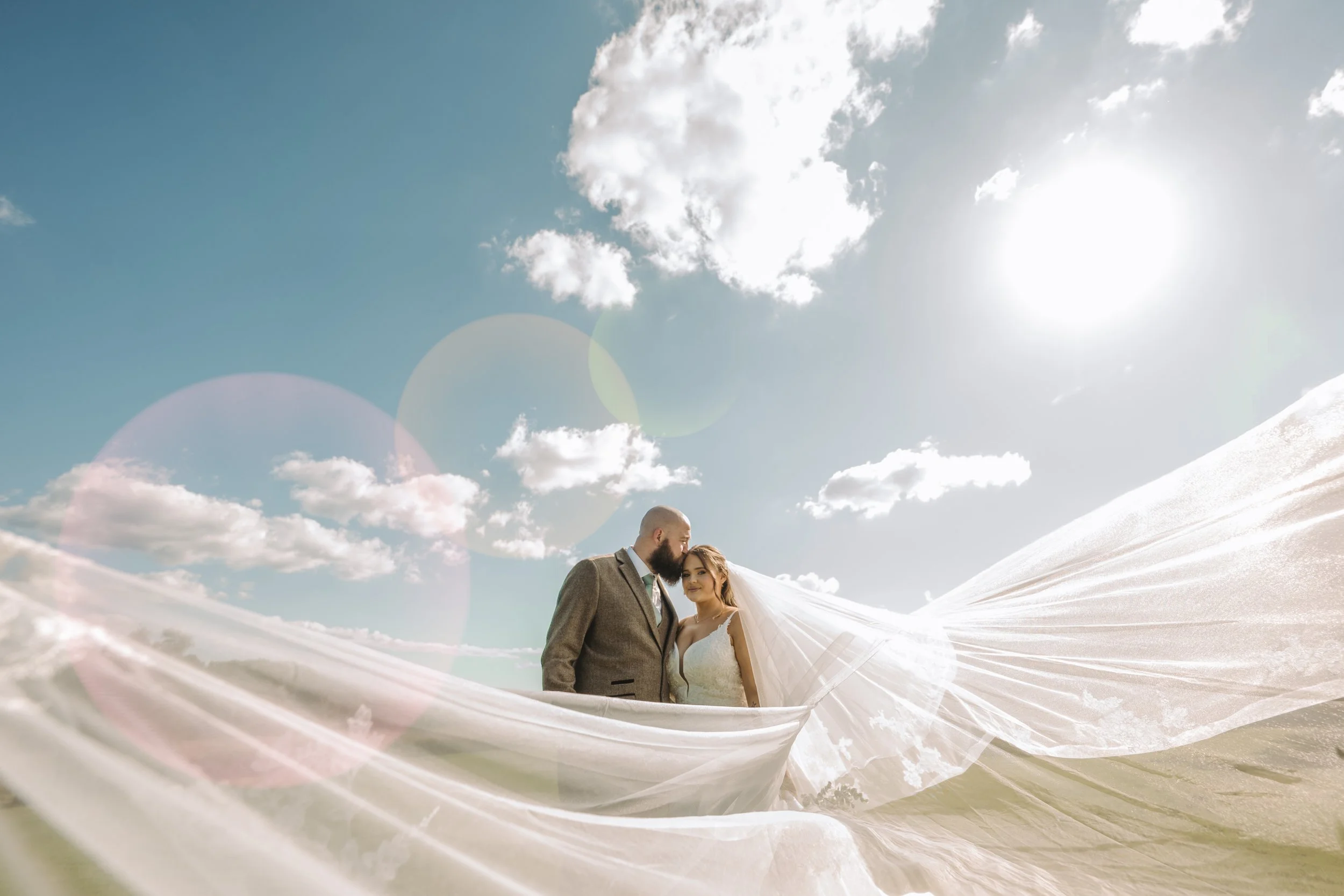 A bride and groom in wedding attire standing outdoors on snow-covered ground under a blue sky with clouds, with the bride's veil flowing in the wind.