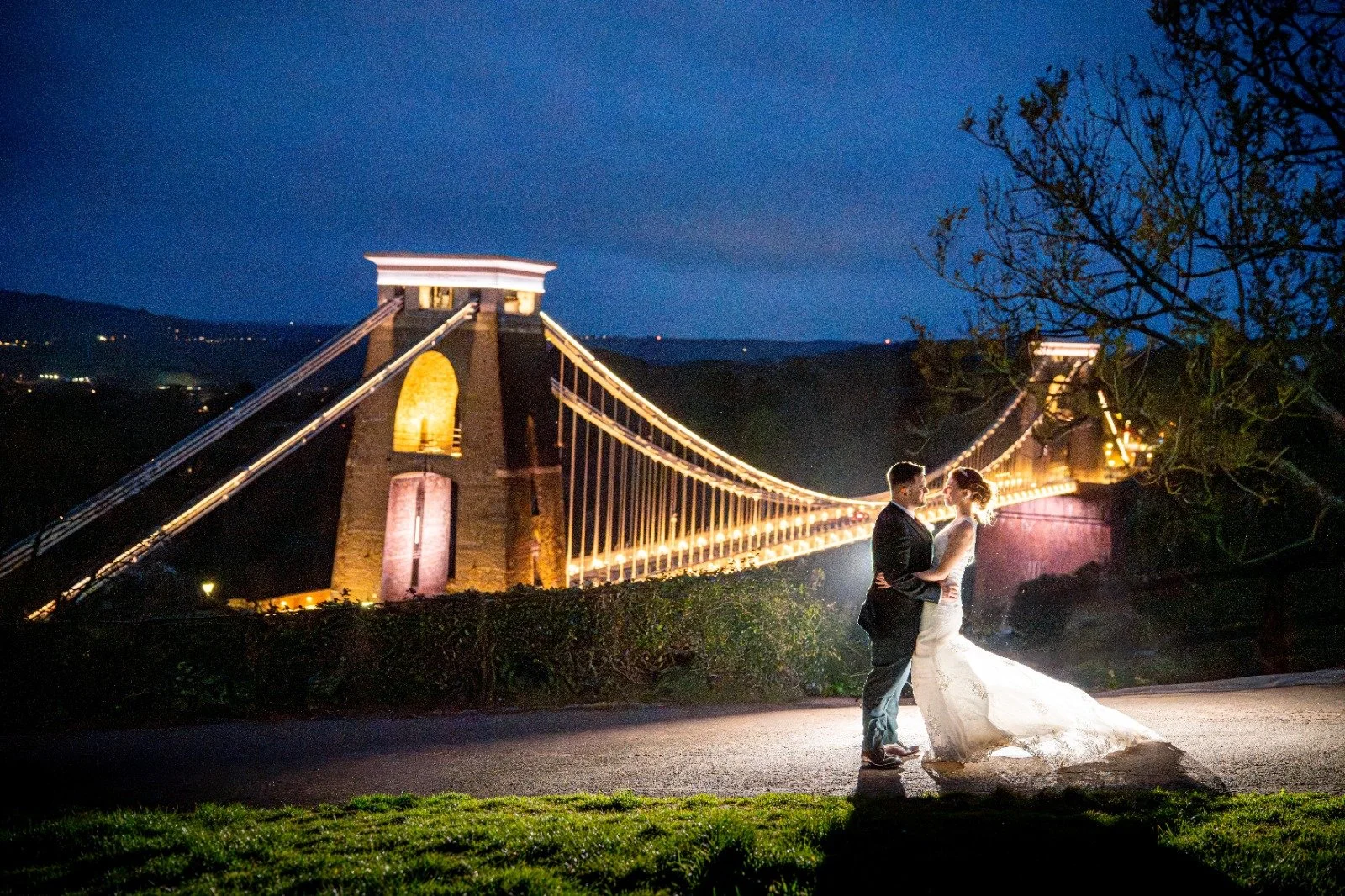 A couple in wedding attire dancing outdoors at night with the illuminated Clifton Suspension Bridge in the background.