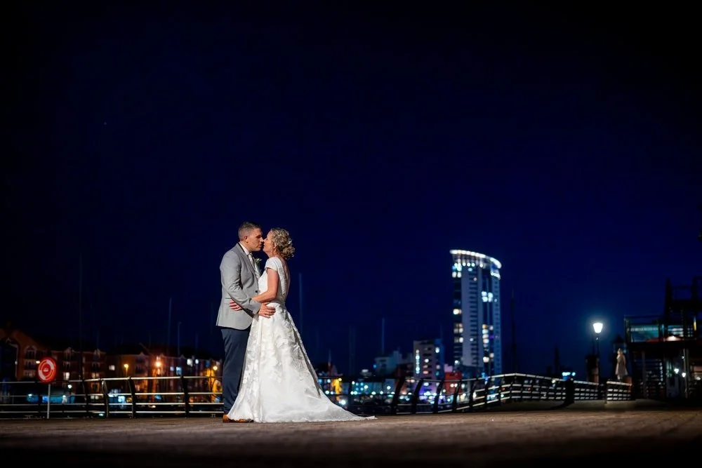 A bride and groom sharing a kiss on a waterfront at night, with city buildings illuminated in the background.