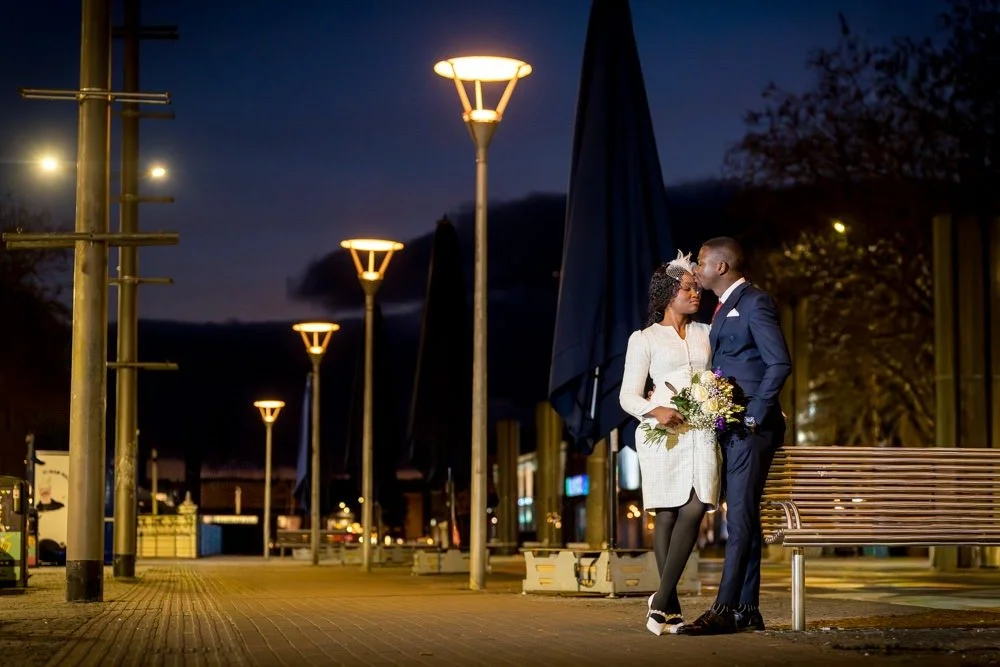A couple in wedding attire sharing a kiss at night on a city sidewalk with street lamps and benches.