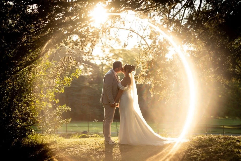 A bride and groom stand in a forest clearing during sunset, holding hands and leaning in for a kiss, with a bright light forming a circle around them.