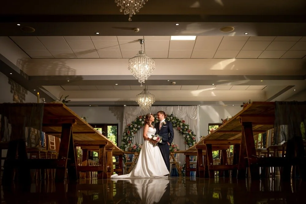A bride and groom standing in a decorated reception hall, gazing at each other. The bride wears a white wedding gown and holds a bouquet, while the groom wears a dark suit. The hall has chandeliers and a floral arch in the background.