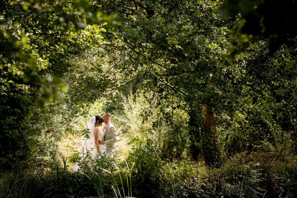 A bride and groom stand close together in a lush, green forest, sharing a tender moment.