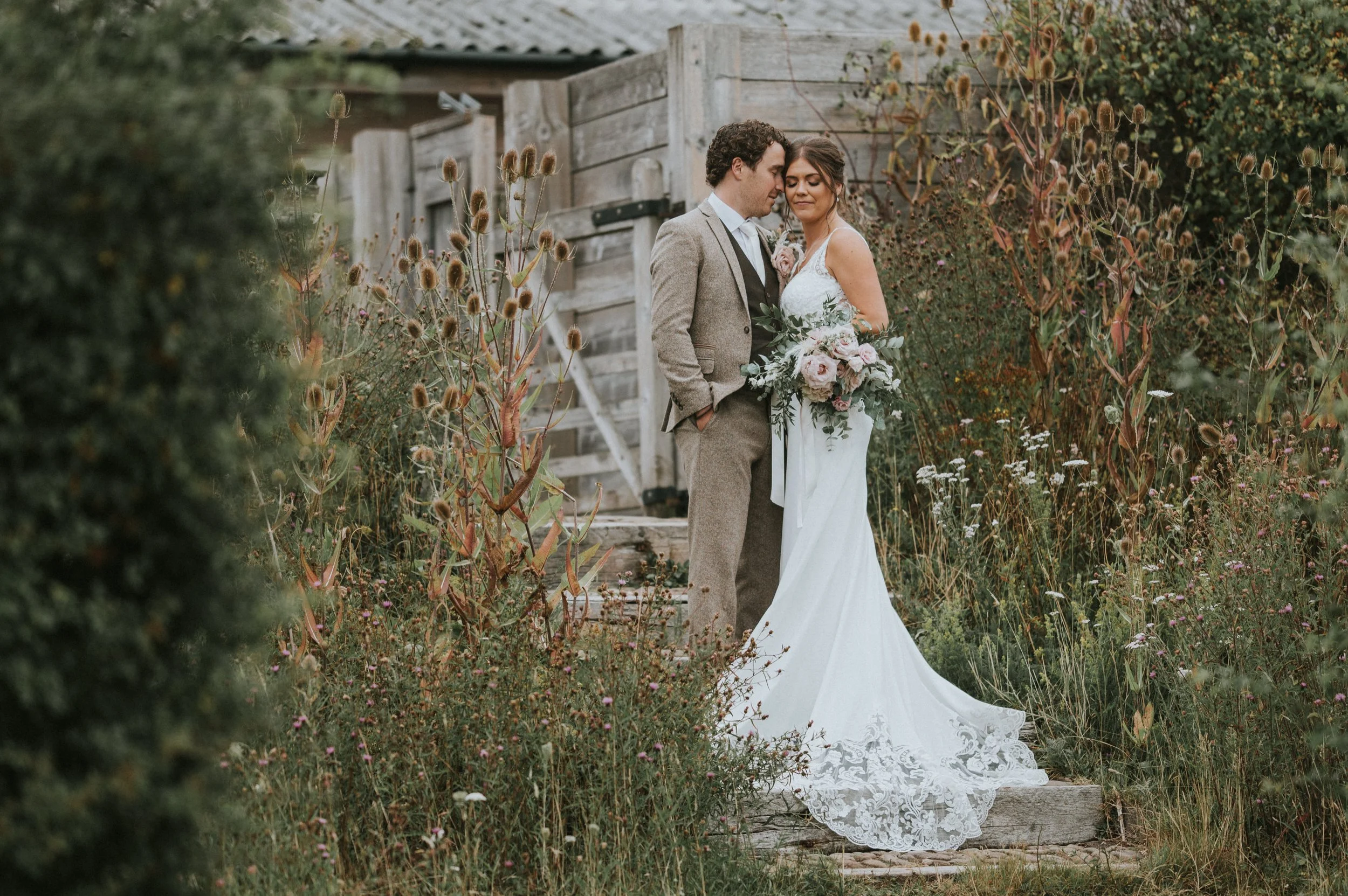A bride and groom standing close together among wildflowers and greenery outdoors, with a rustic wooden fence behind them, on their wedding day.
