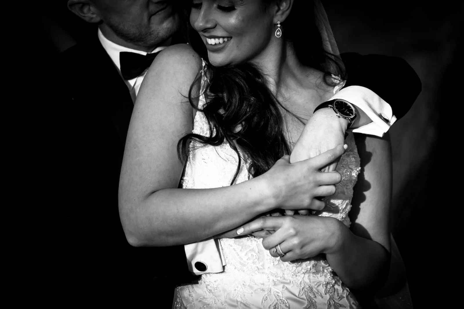 A black and white photo of a smiling bride and groom embracing. The bride is wearing a wedding dress with lace details, and the groom is in a tuxedo with a bow tie. The bride is showing her wedding ring, and the groom is wearing a watch on his wrist.
