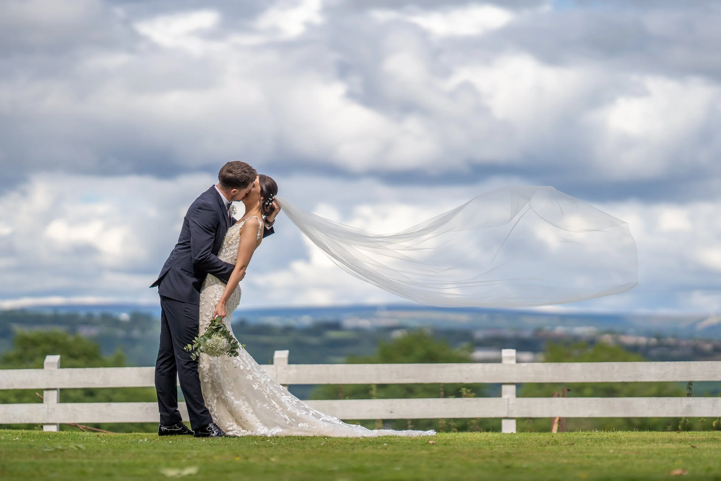 A bride and groom sharing a kiss outdoors on a cloudy day, with the bride holding a bouquet of white flowers and wearing a long lace wedding dress, and the groom in a dark suit, standing near a white wooden fence with a scenic landscape in the backgr