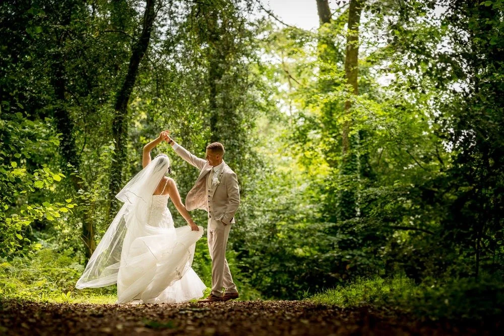 A bride and groom dancing in a lush green forest, with the bride wearing a white wedding dress and veil, and the groom in a light-colored suit.