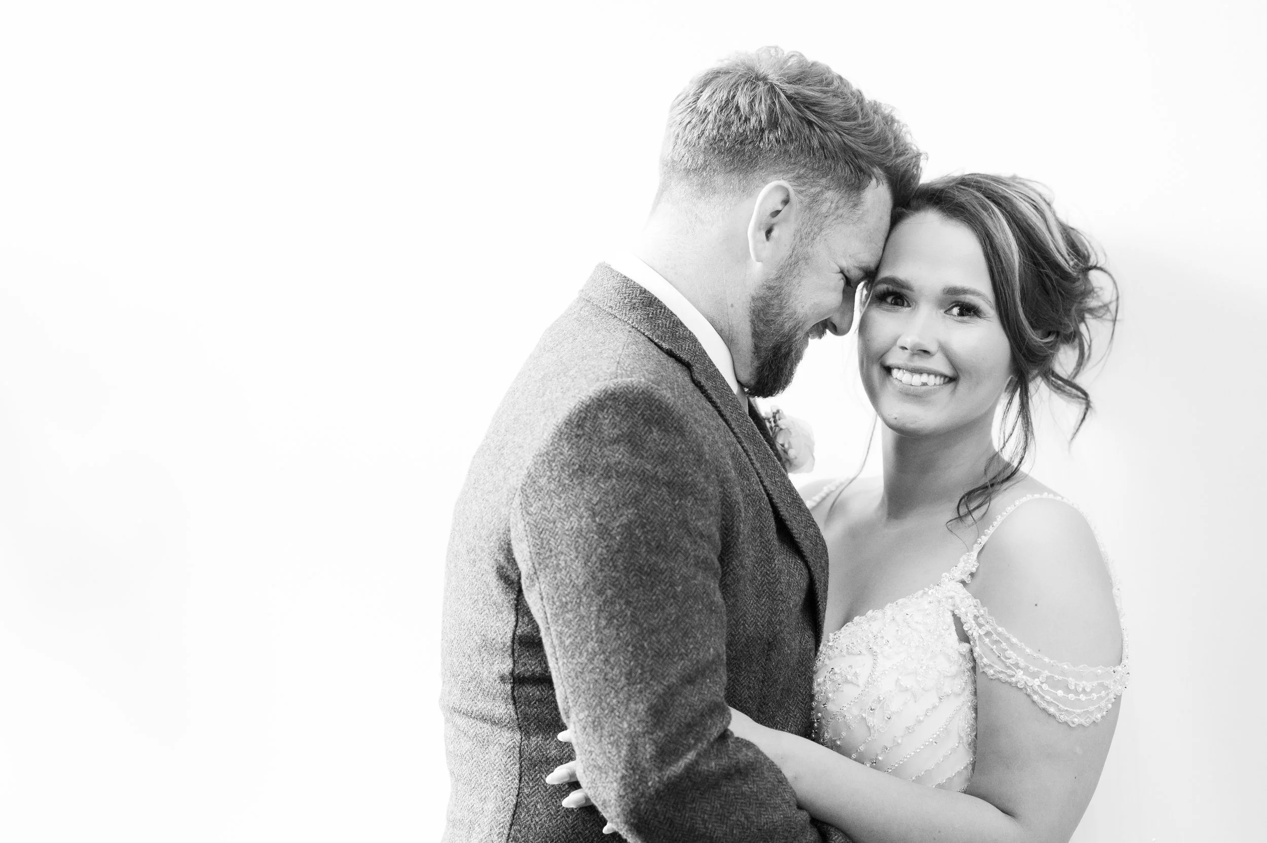 A black-and-white photo of a couple on their wedding day. The man has a beard and is wearing a suit, and the woman has curly hair and is wearing a wedding dress. They are smiling and standing close together, with their foreheads touching and eyes clo