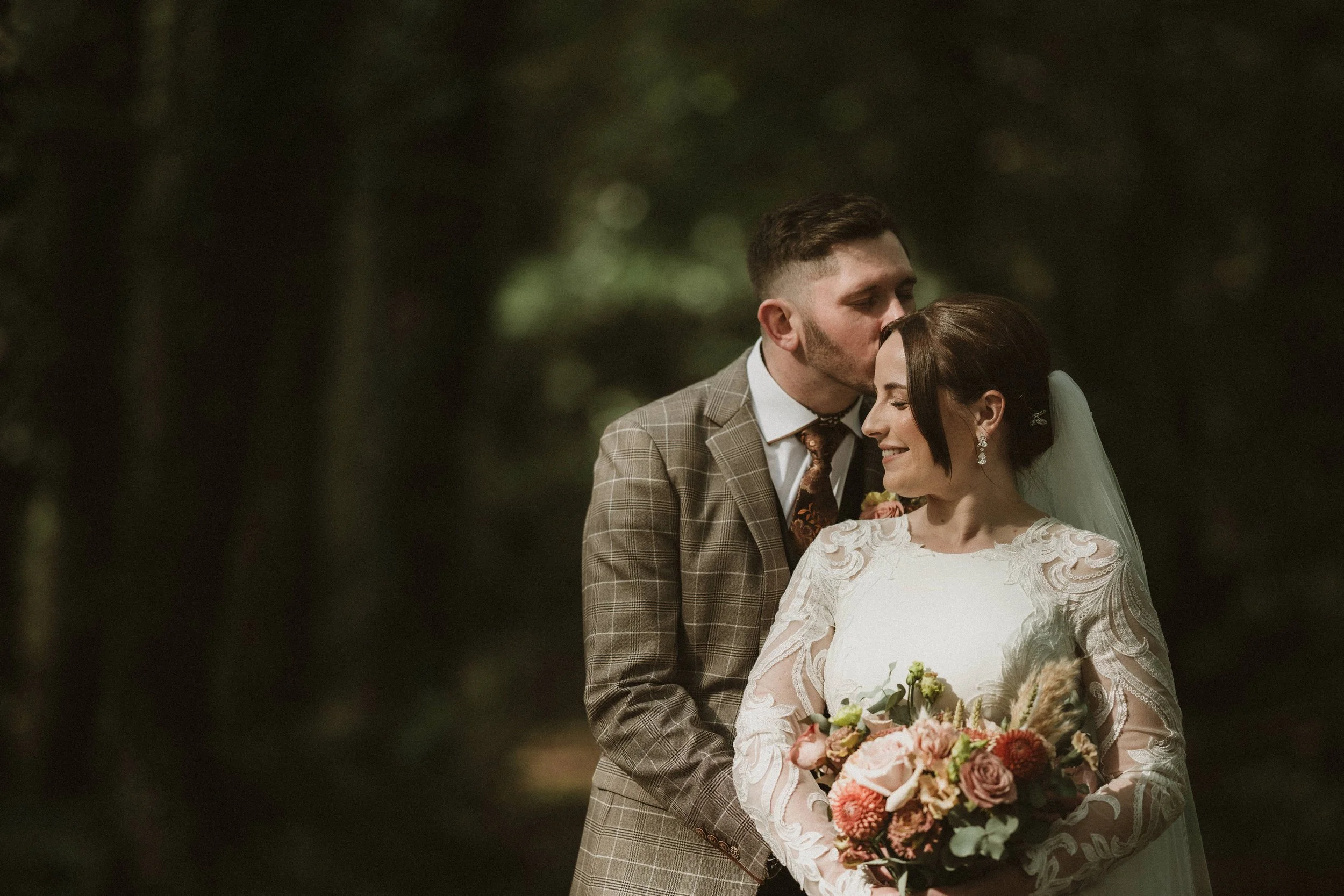 A groom wearing a checked suit is kissing a bride on her forehead during a wedding ceremony. The bride is holding a bouquet of flowers and is dressed in a lace wedding gown. They are standing outdoors in a natural setting at night.