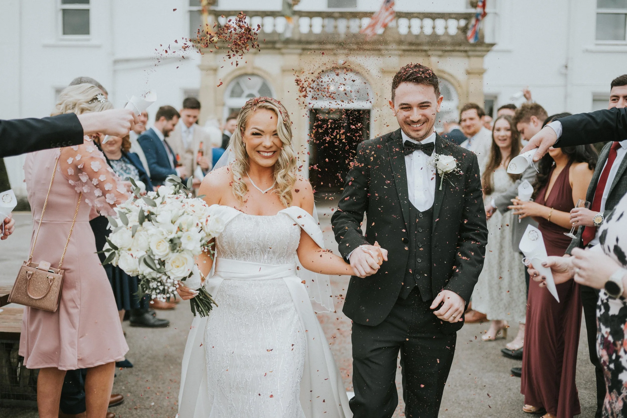A newly married couple smiling and holding hands as friends celebrate by tossing confetti outside a historic building.