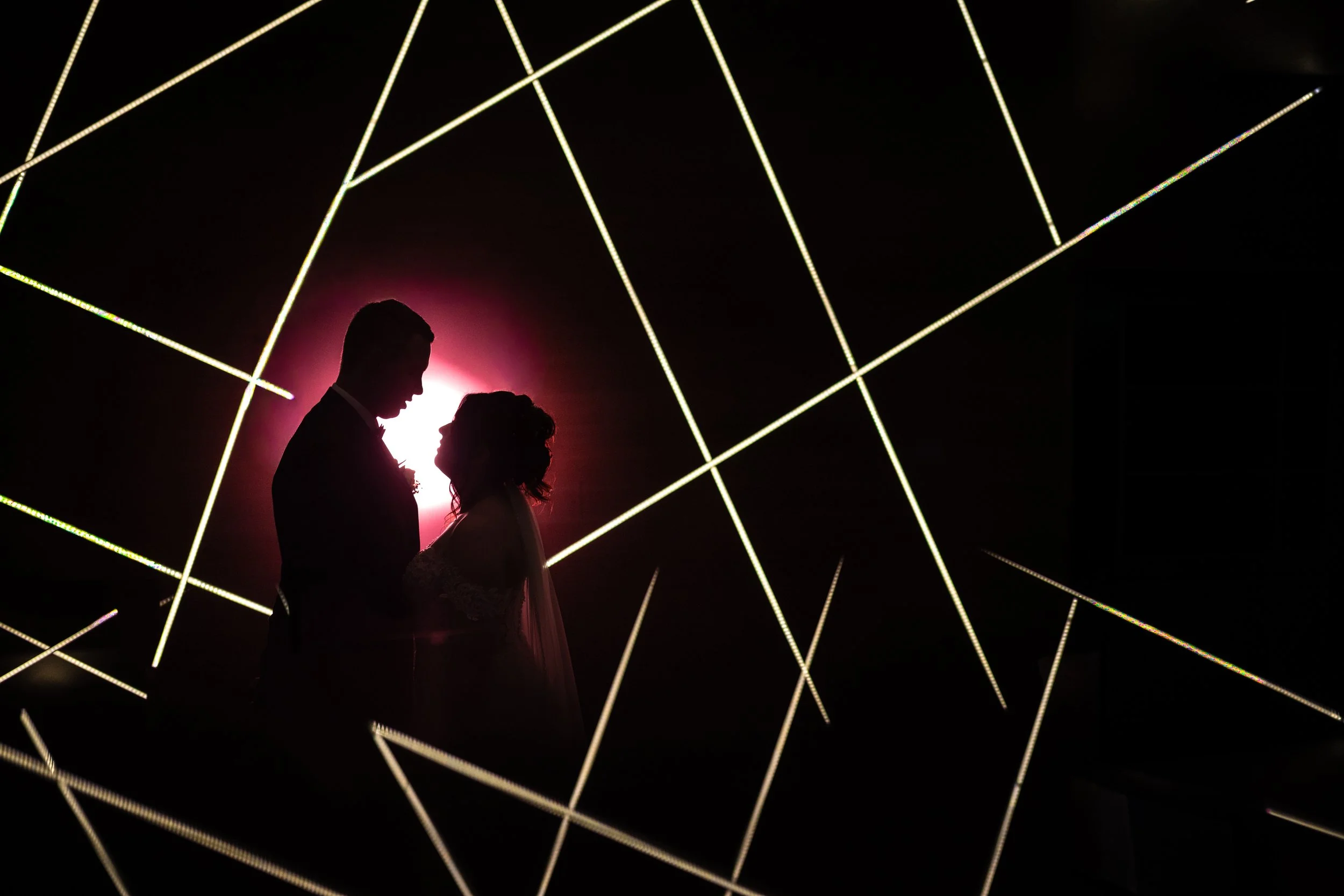 Silhouette of a couple, a man and woman, facing each other against a pink light, with decorative light beams in the background.