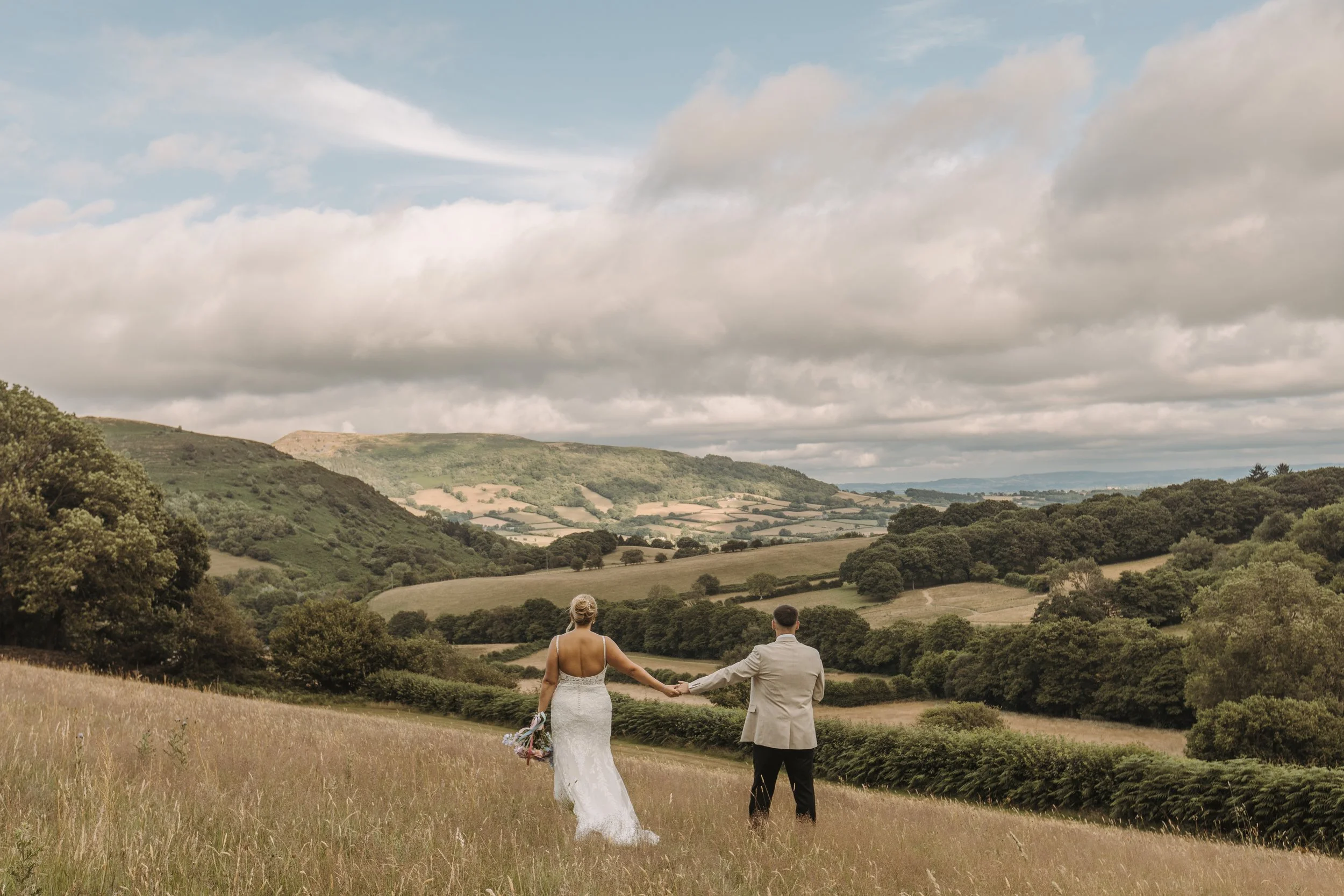 A bride and groom holding hands, walking through a grassy field towards rolling hills under a partly cloudy sky.