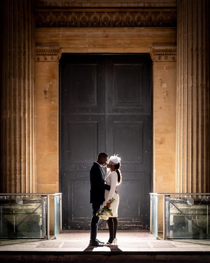 A bride and groom standing close together in front of a large dark door, illuminated by backlighting, with the bride holding a bouquet of flowers.