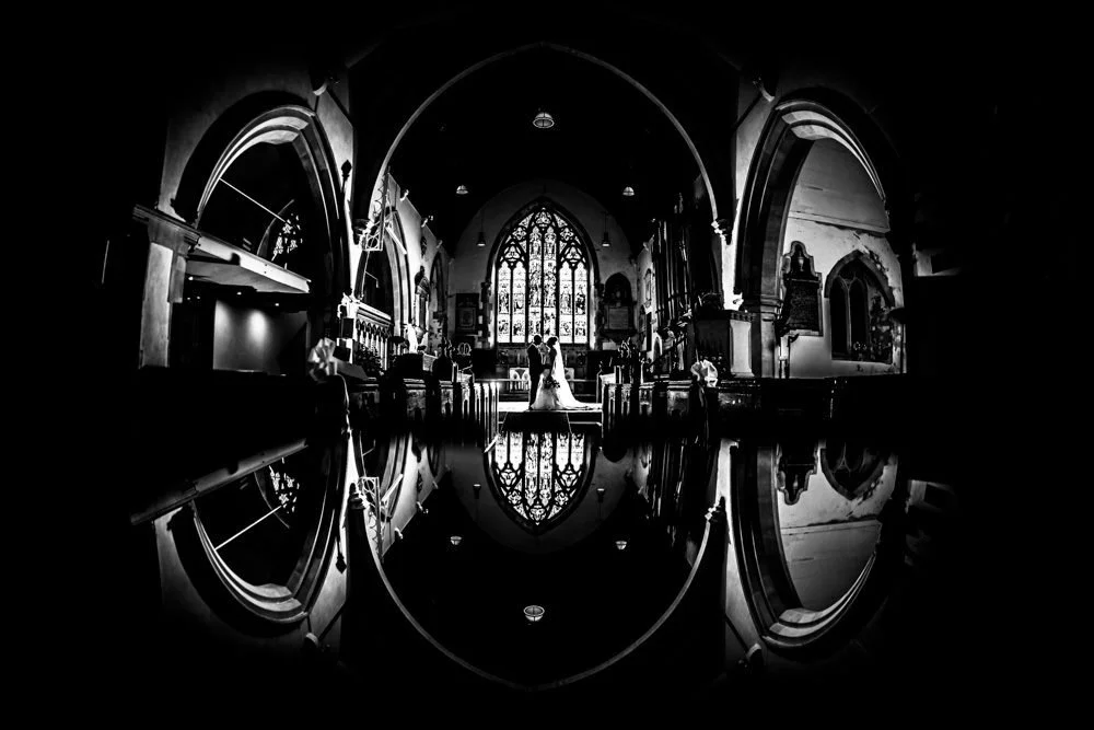 Black and white photo of a church interior with a couple at the altar, stained glass windows in the background, and reflections on the floor creating an oval frame around the scene.