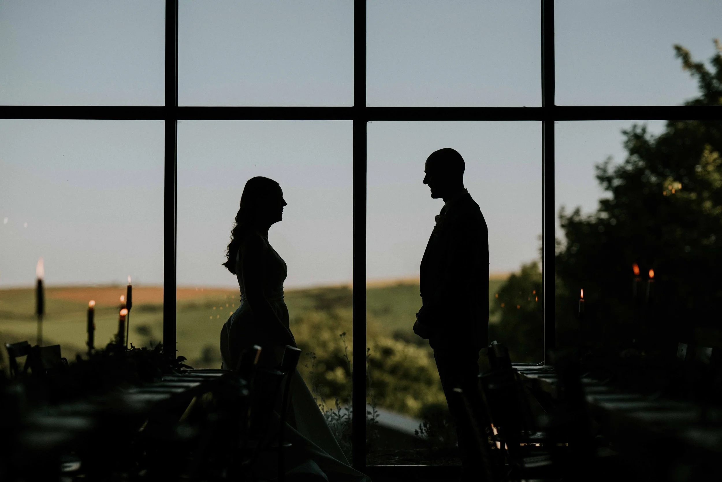 Silhouettes of a woman and a man facing each other inside a building with large window panes, with a cloudy sky and trees visible outside.