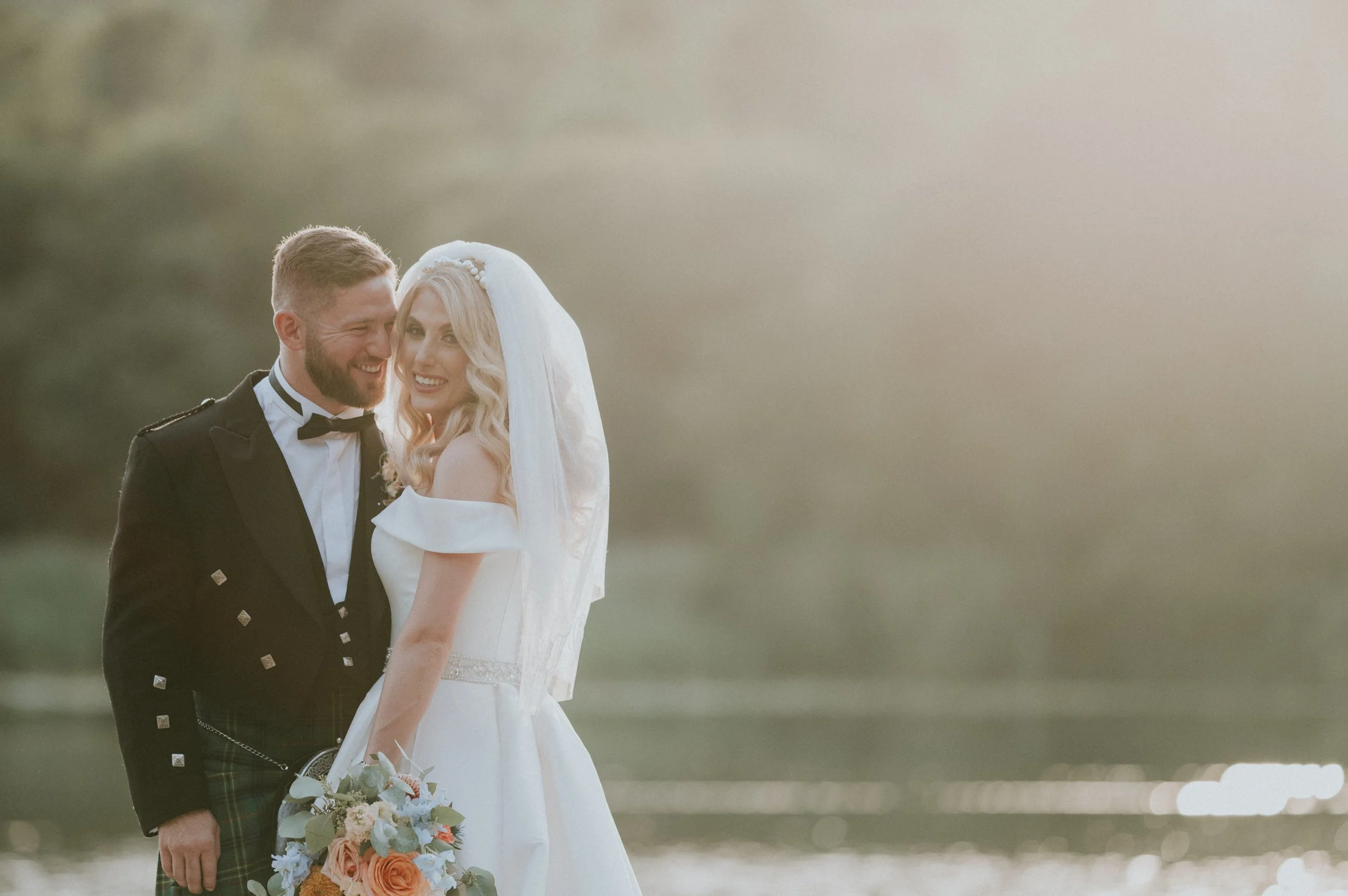 A newlywed couple smiling and close together outdoors, the groom in a black tuxedo with a bow tie, the bride in a white wedding gown with a veil, holding a bouquet of flowers, with a blurred natural landscape in the background.