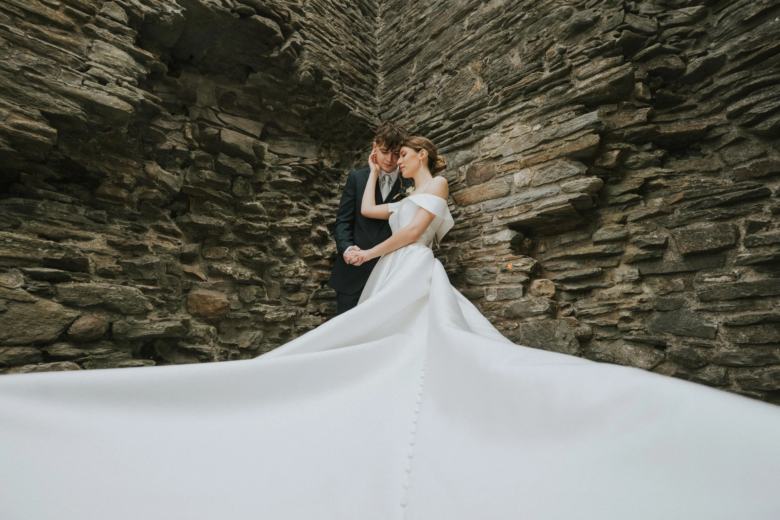 A bride and groom standing close together, holding hands, with a stone wall in the background.