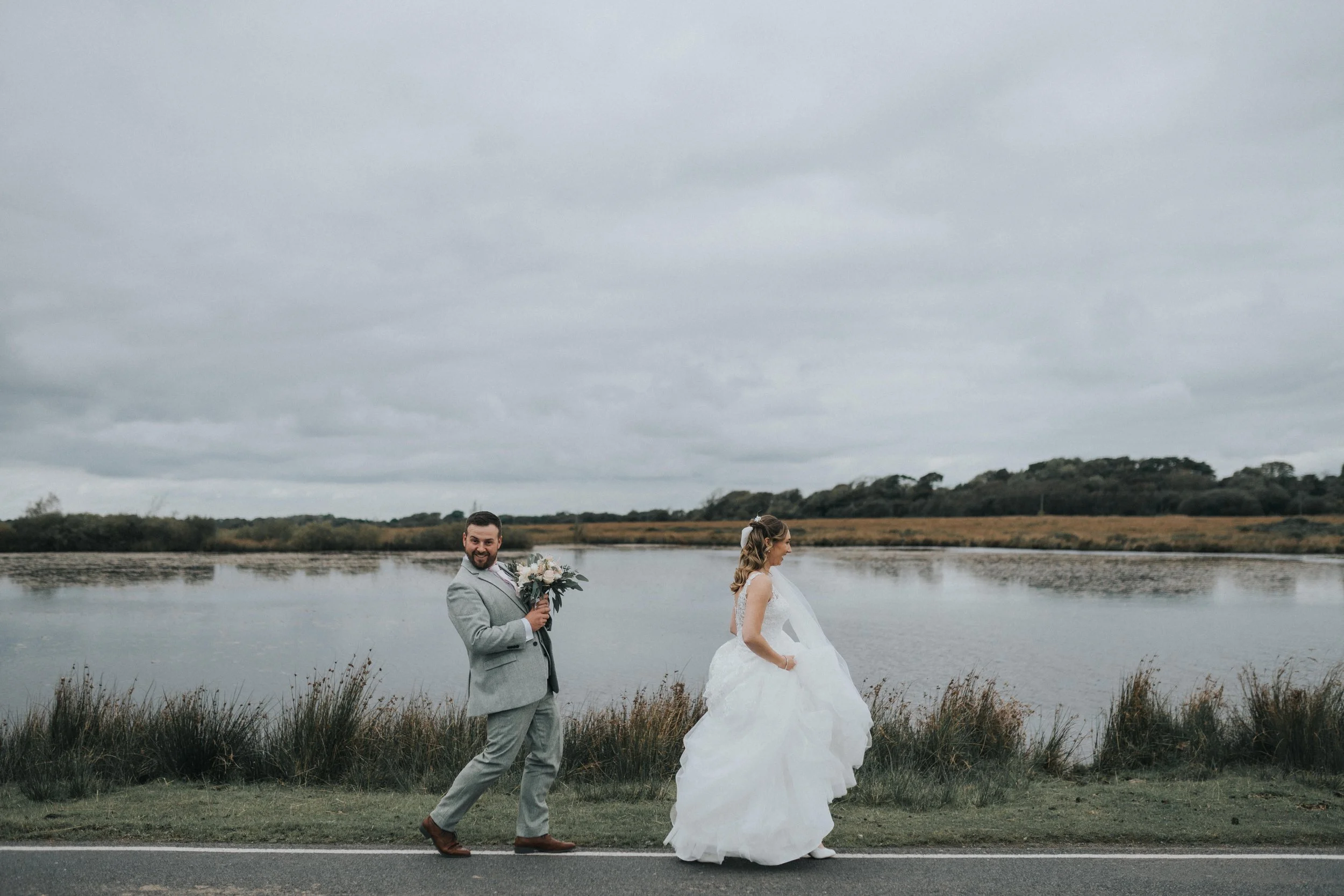 A bride and groom standing outdoors by a lake on an overcast day, with the groom holding a bouquet of flowers and smiling, and the bride facing away from the camera in a white wedding dress.