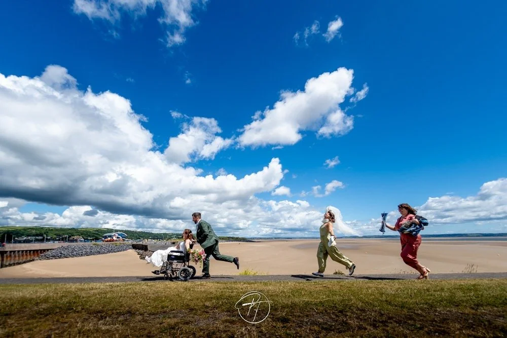 A wedding scene with a bride in a wheelchair and groom running toward her, three other women walking along a beach under a blue sky with clouds.