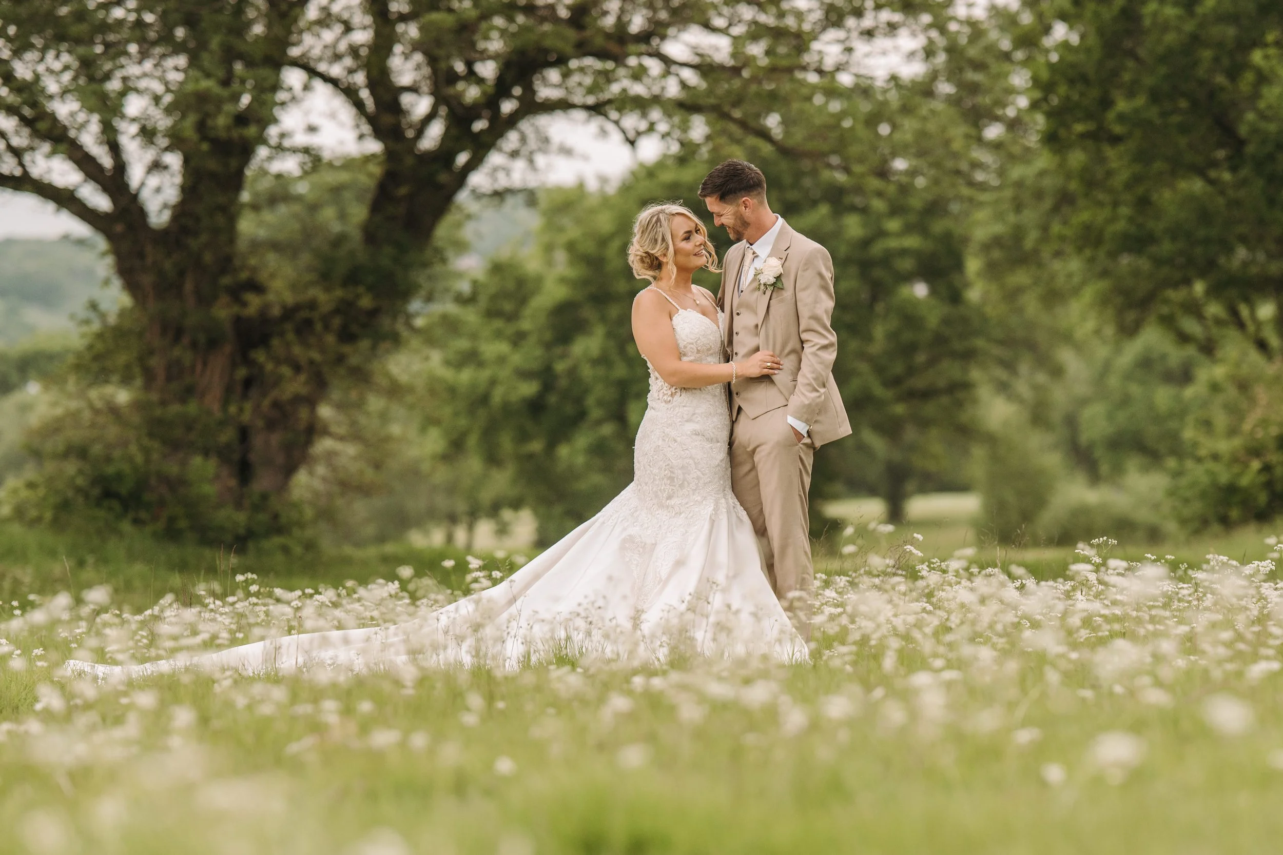 A bride and groom standing close together outdoors in a grassy area with trees in the background, sharing a loving moment.