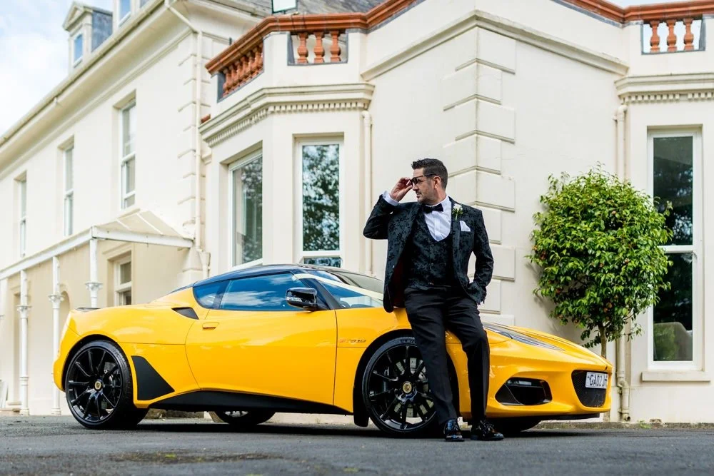A man in a black tuxedo leaning against a yellow sports car in front of a white building with large windows and a small tree.