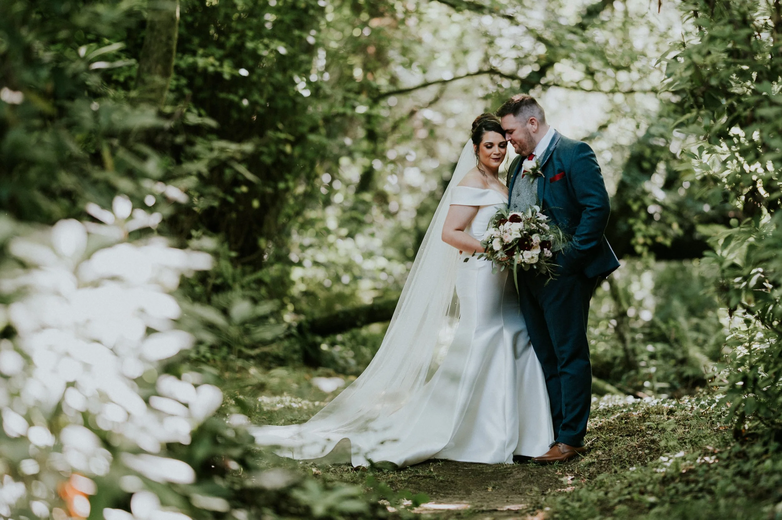 A bride and groom standing close together outdoors among trees, with the bride holding a bouquet of flowers, sharing a tender moment.