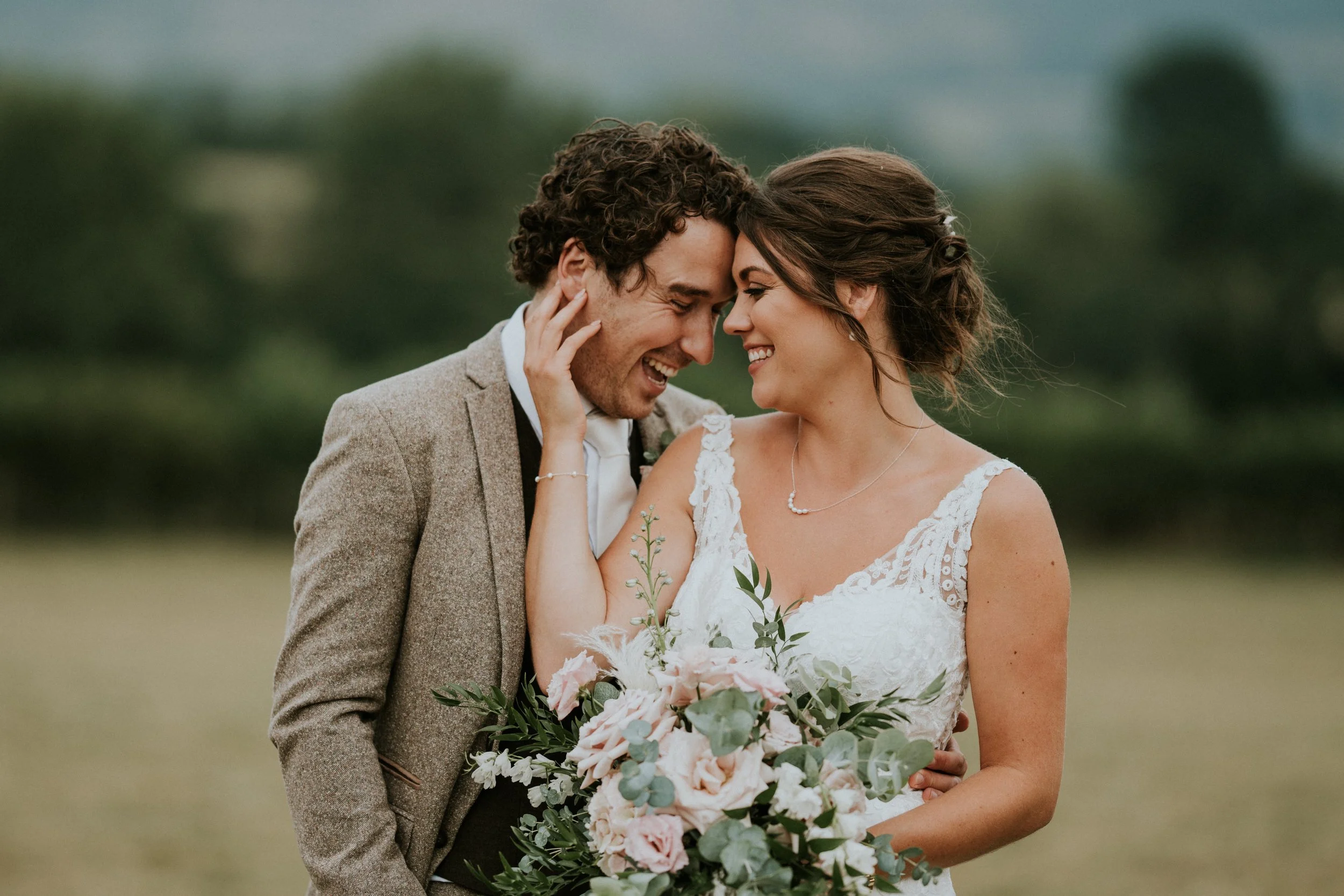 A smiling bride and groom are embracing each other outdoors, with their foreheads touching. The bride is holding a large bouquet of pink and white flowers with greenery. The groom is wearing a beige suit with a white shirt, and the bride is wearing a