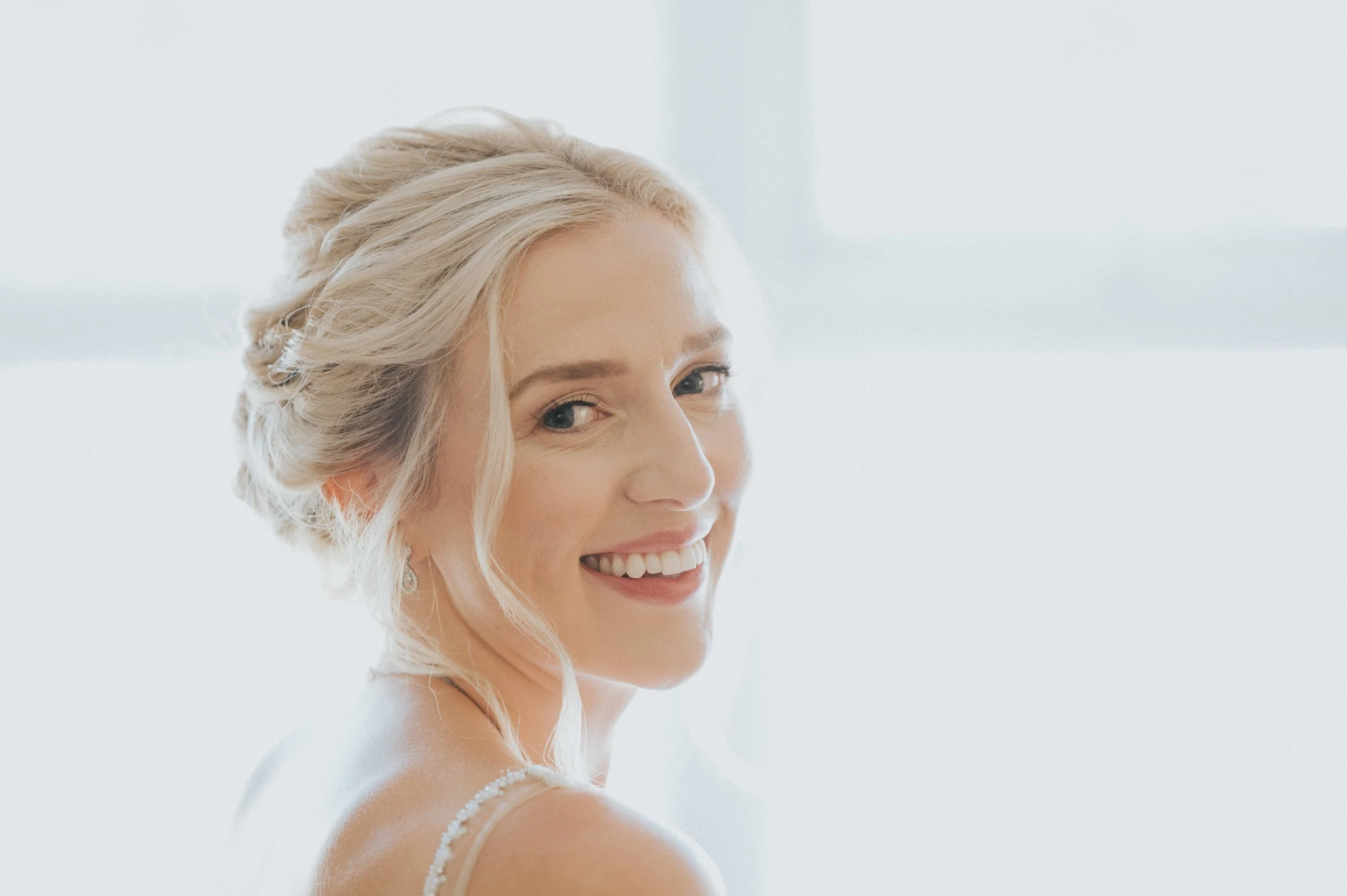 Close-up of a smiling woman with blonde hair styled in an elegant updo, wearing a white dress with thin straps, against a bright, minimalistic background.