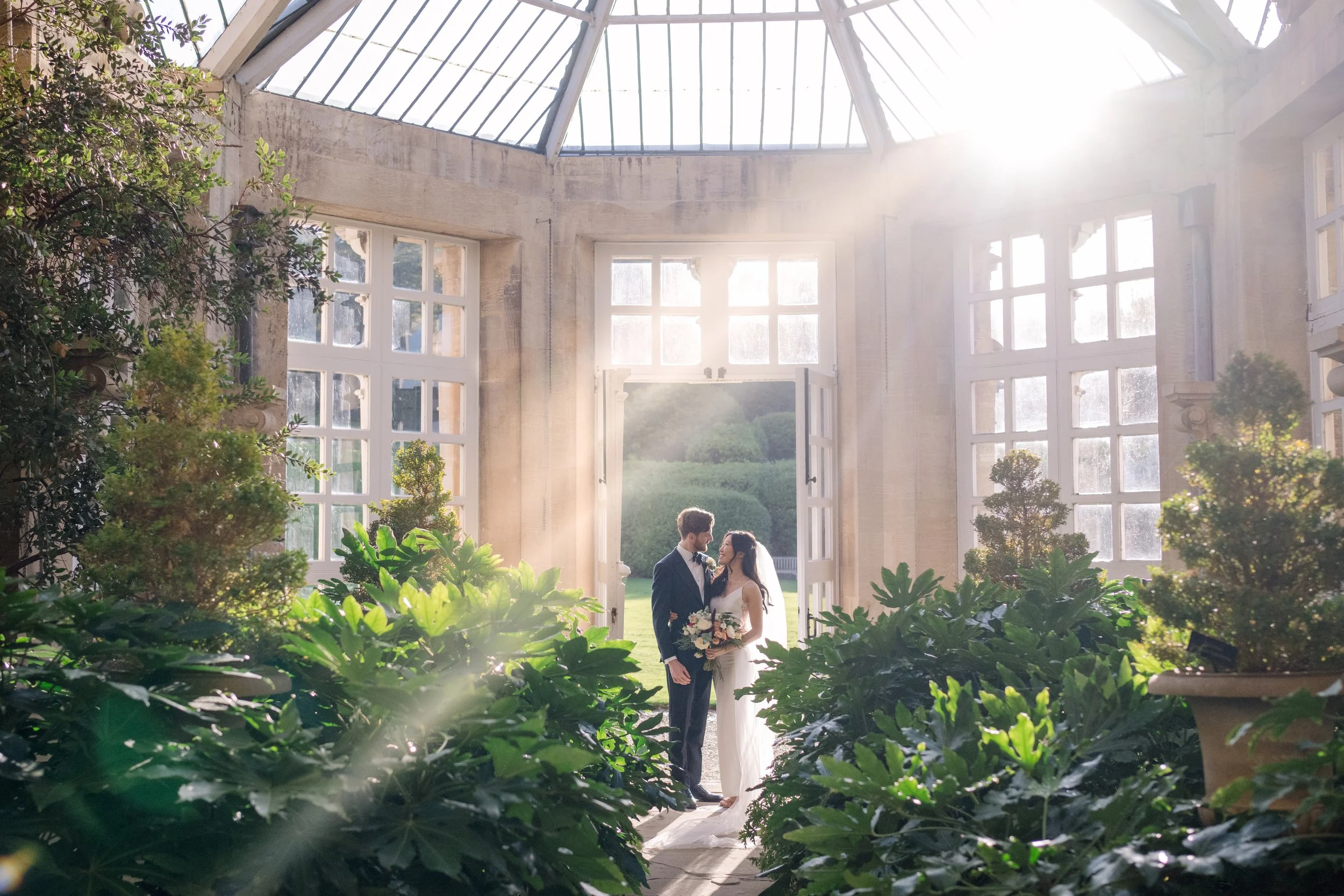 A bride and groom standing together in a bright greenhouse surrounded by lush green plants, with sunlight streaming through large windows and roof, creating a romantic and airy atmosphere.