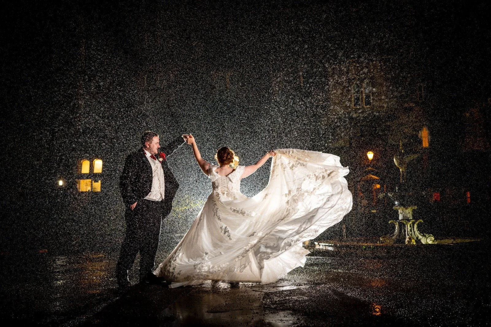 Nighttime wedding scene with a bride and groom dancing amid falling water droplets, illuminated by surrounding lights; bride wears a flowing white wedding dress, groom in a tuxedo.